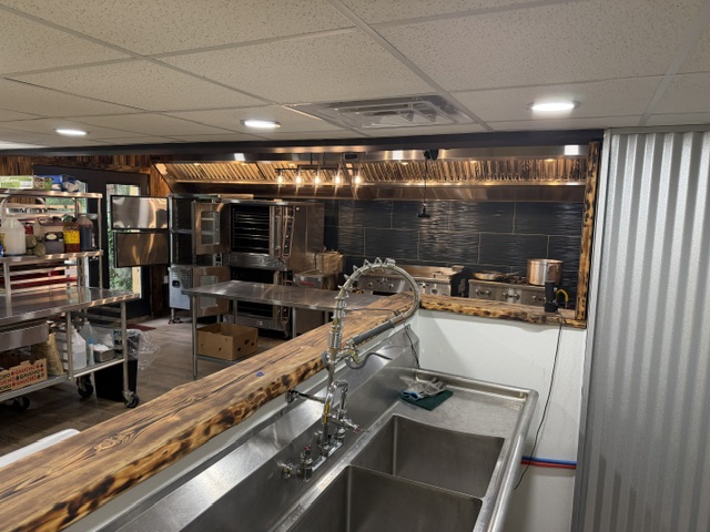 Commercial kitchen with stainless steel appliances, wooden countertop, and cooking equipment, viewed from the side.