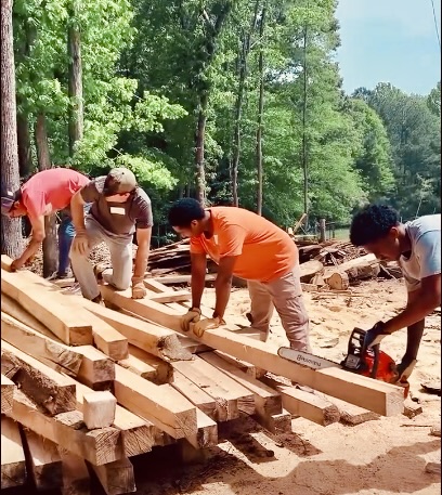Four people working outdoors on a wooden construction project in a forested area, with trees and logs in the background.