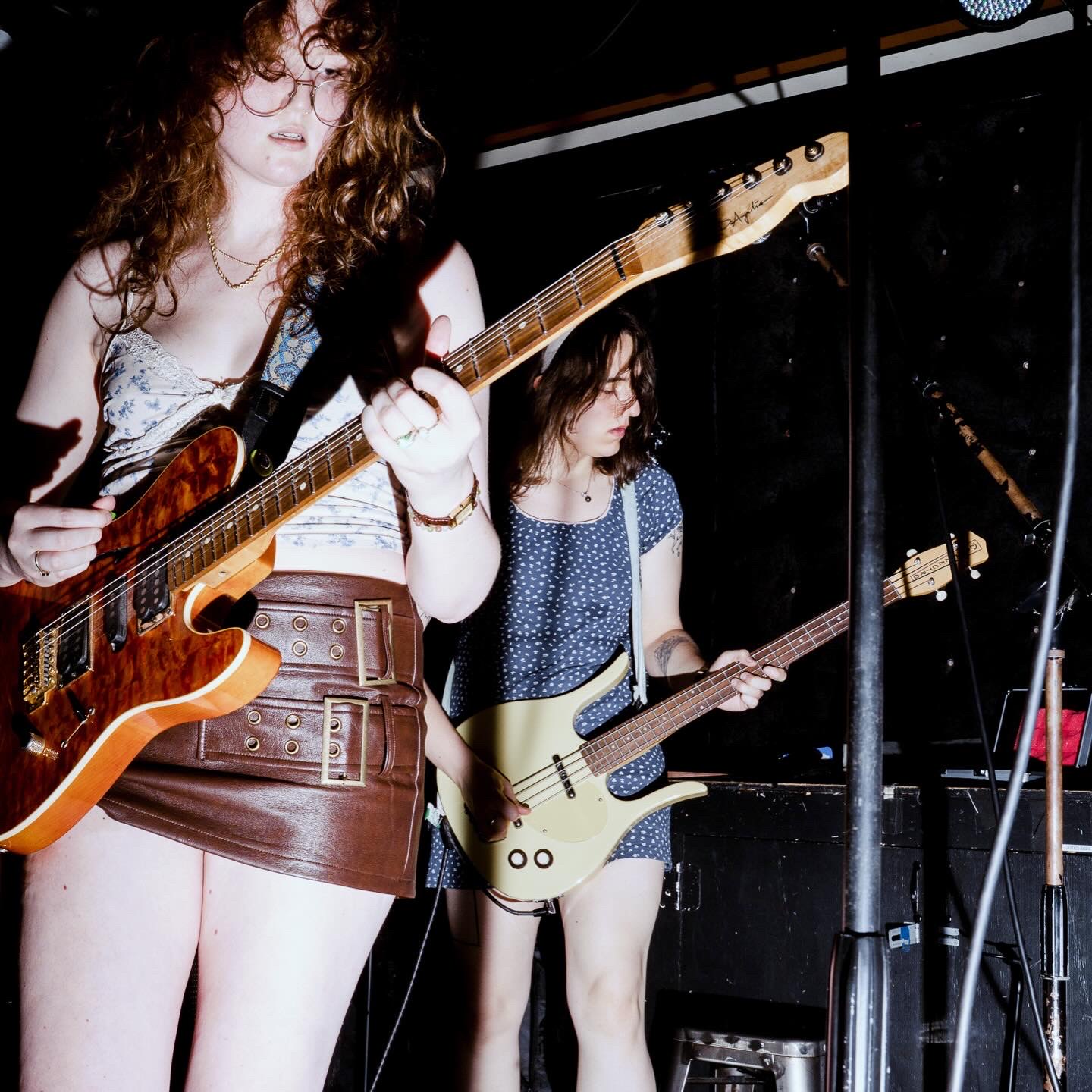 Two young women playing electric guitars on stage, one with curly hair and glasses, the other with straight hair, in a dark setting.