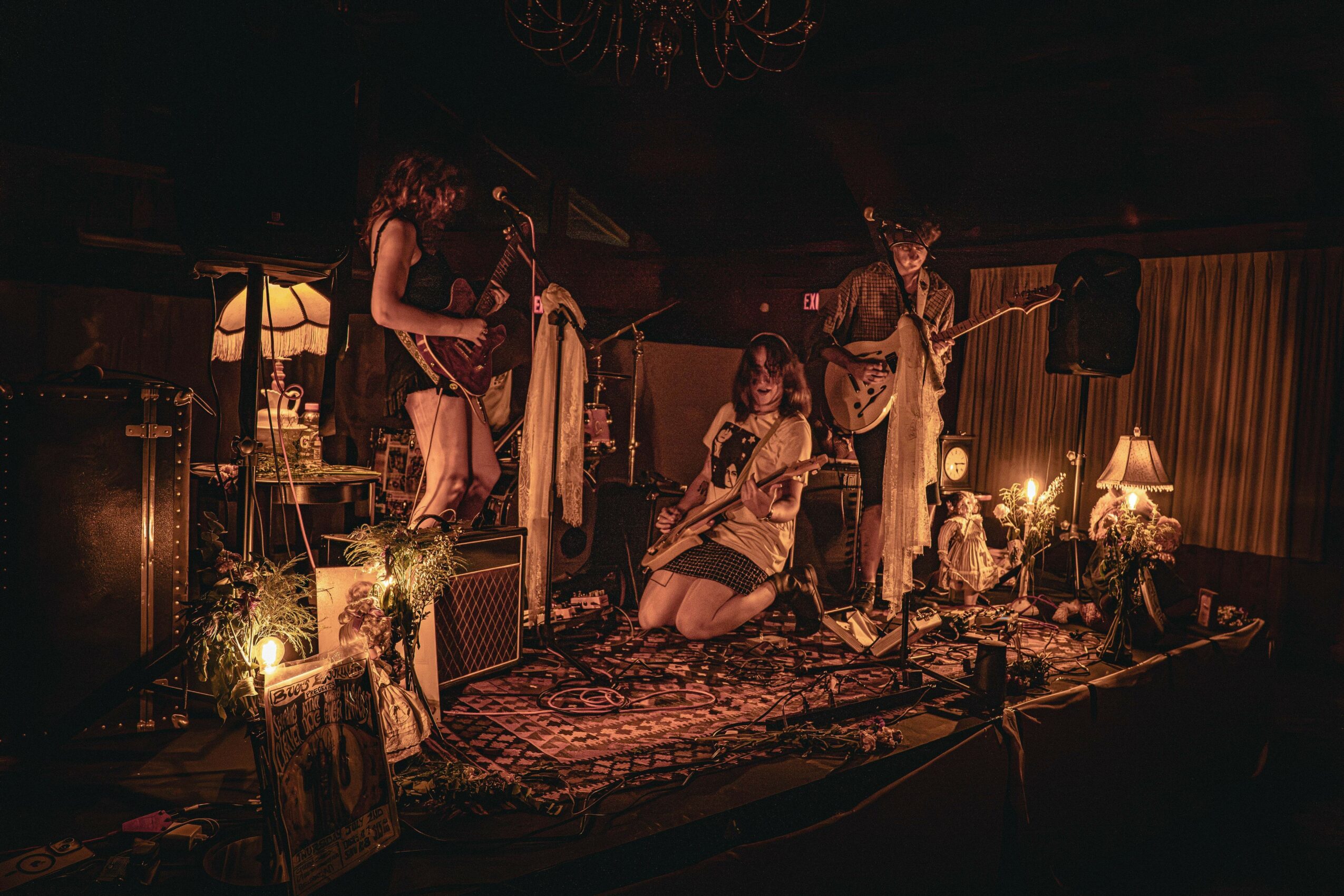 Three women perform on a dimly lit stage with musical instruments and vintage decor, including lamps and framed pictures.