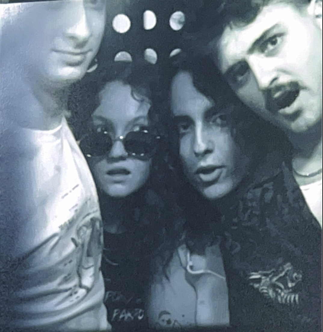 Four people close together, two women and two men, in black and white, with one woman wearing sunglasses, all looking at the camera.