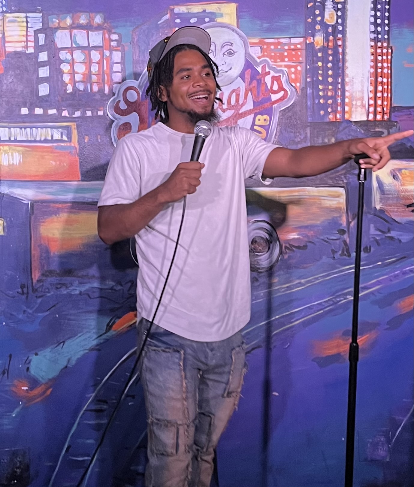 Person with headphones holding a microphone, smiling, pointing, standing in front of a colorful cityscape mural.