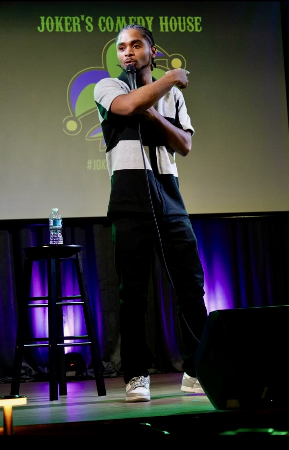 Stand-up comedian on stage with a microphone, a stool with water, and a screen behind displaying 'Joker's Comedy House'.