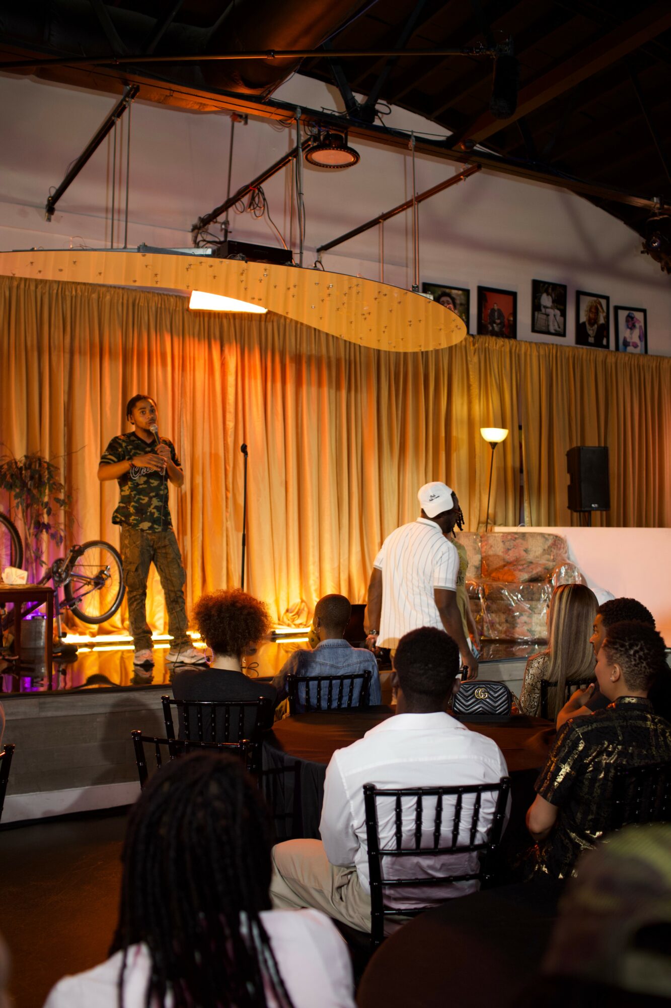 Person on stage speaking to audience in a venue with golden curtains and framed pictures on wall, audience seated and listening.