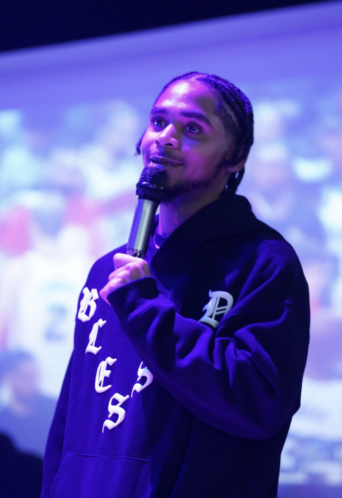 Person with braided hair holding a microphone, wearing a dark hoodie, on stage with a blurred background.