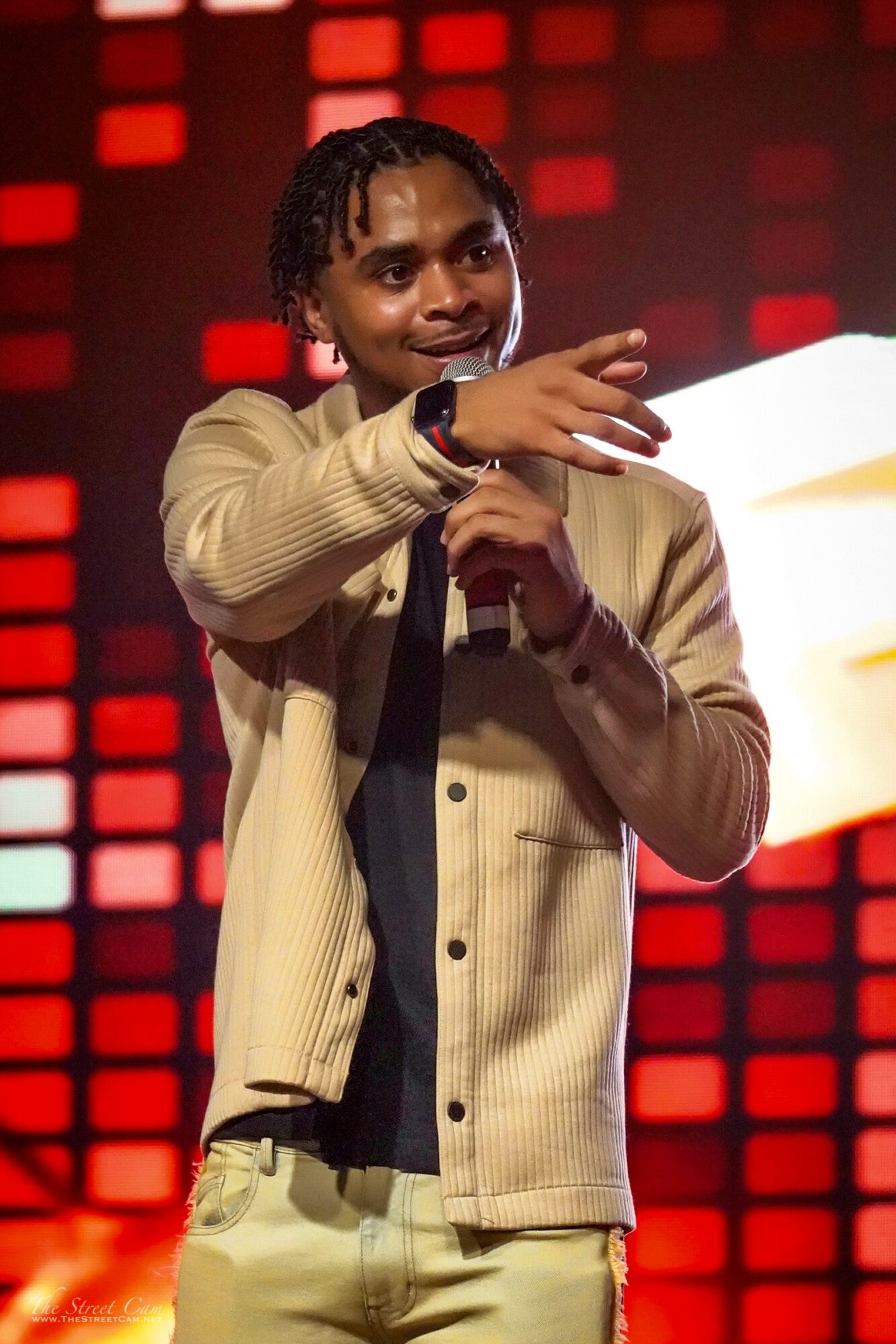 Person holding microphone and gesturing with hand on stage with red and black background, smiling.