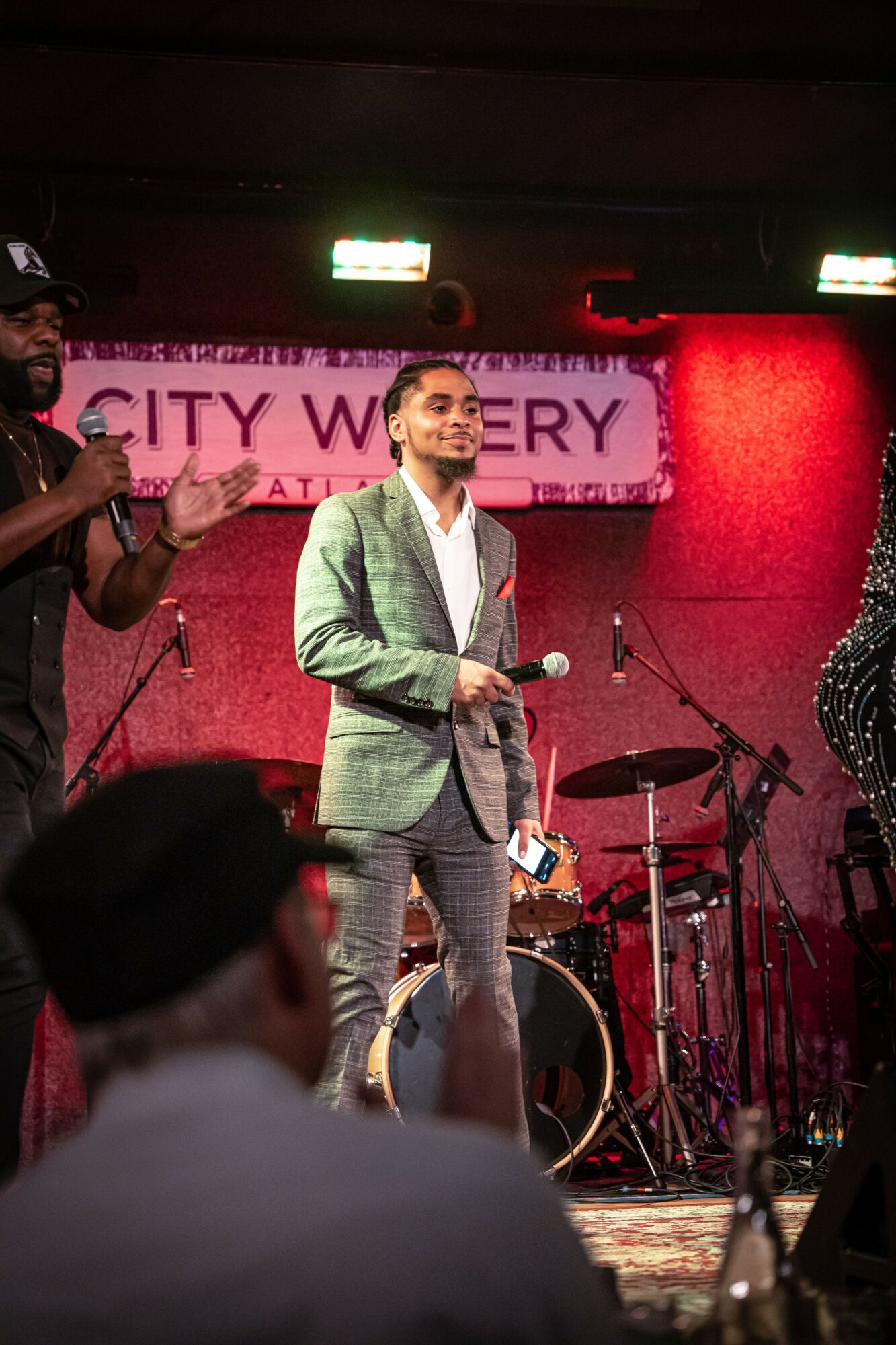 Person in a gray suit holding a microphone on stage with a band and a pink sign in the background.