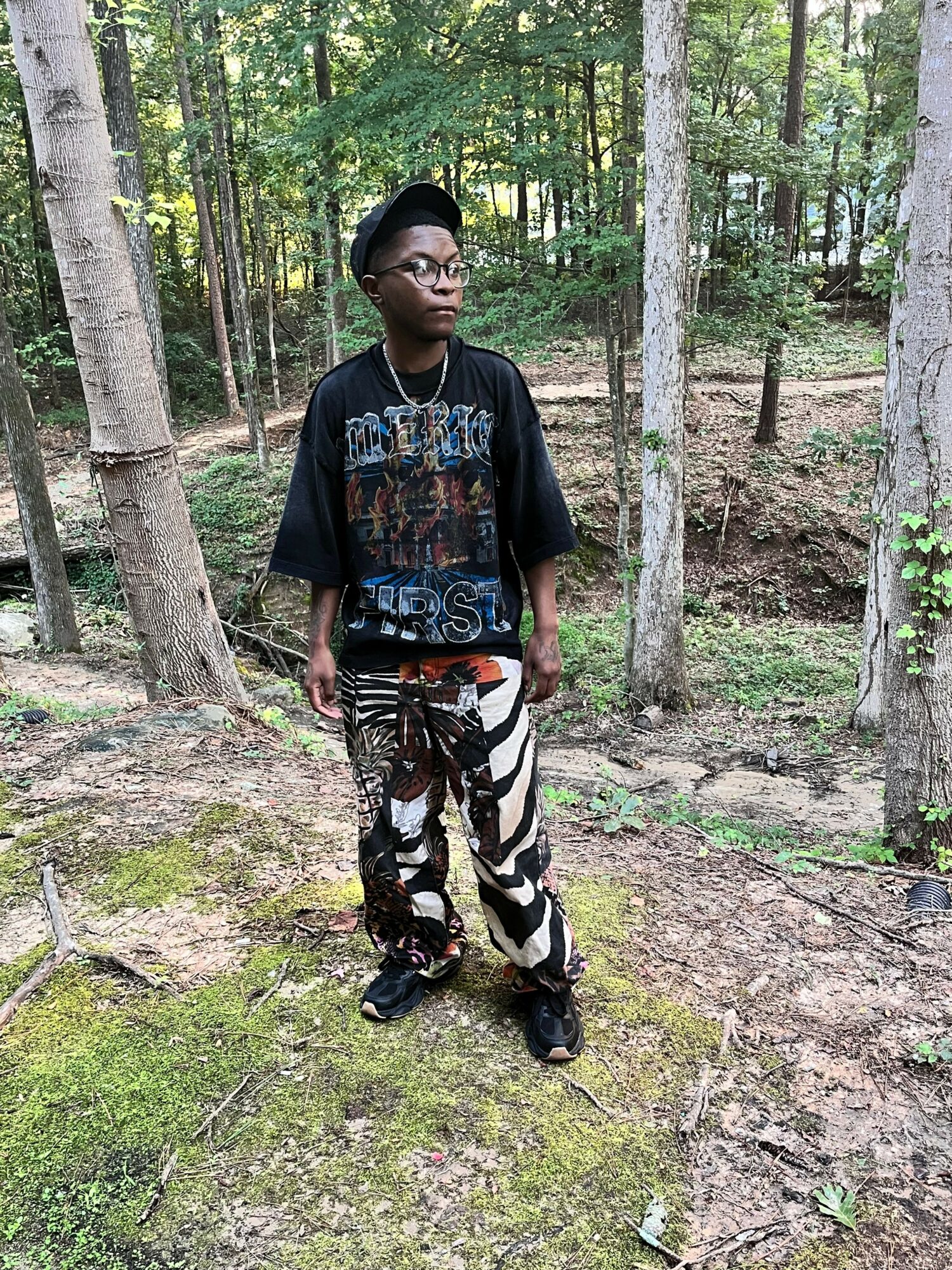 Person standing in a forest with trees and mossy ground, holding a walking stick, wearing sunglasses, a graphic t-shirt, and camouflage pants.
