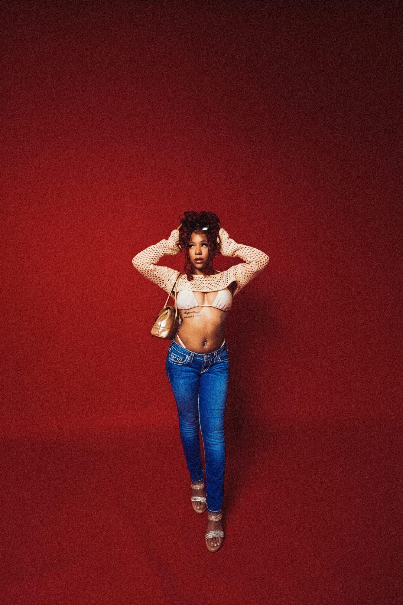 Woman standing with hands behind head, wearing a white crop top and blue jeans, against a red background.