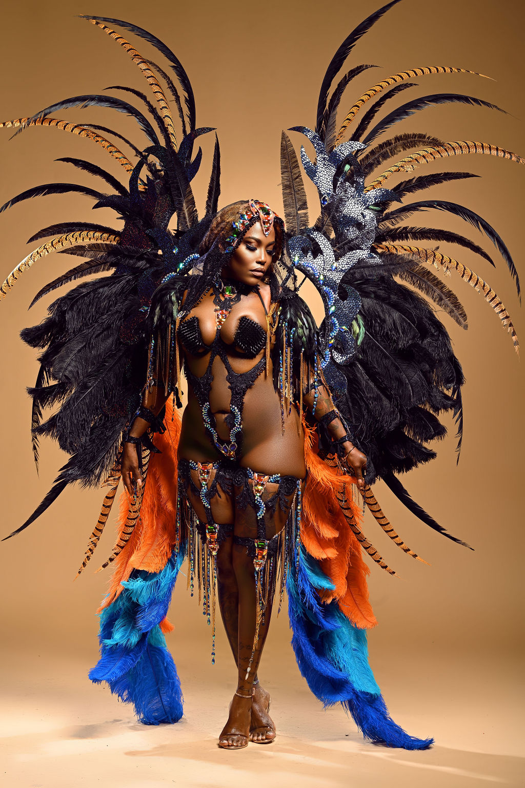 Person in elaborate costume with large black feathered wings, colorful fabric, and decorative accessories, standing against a plain background.