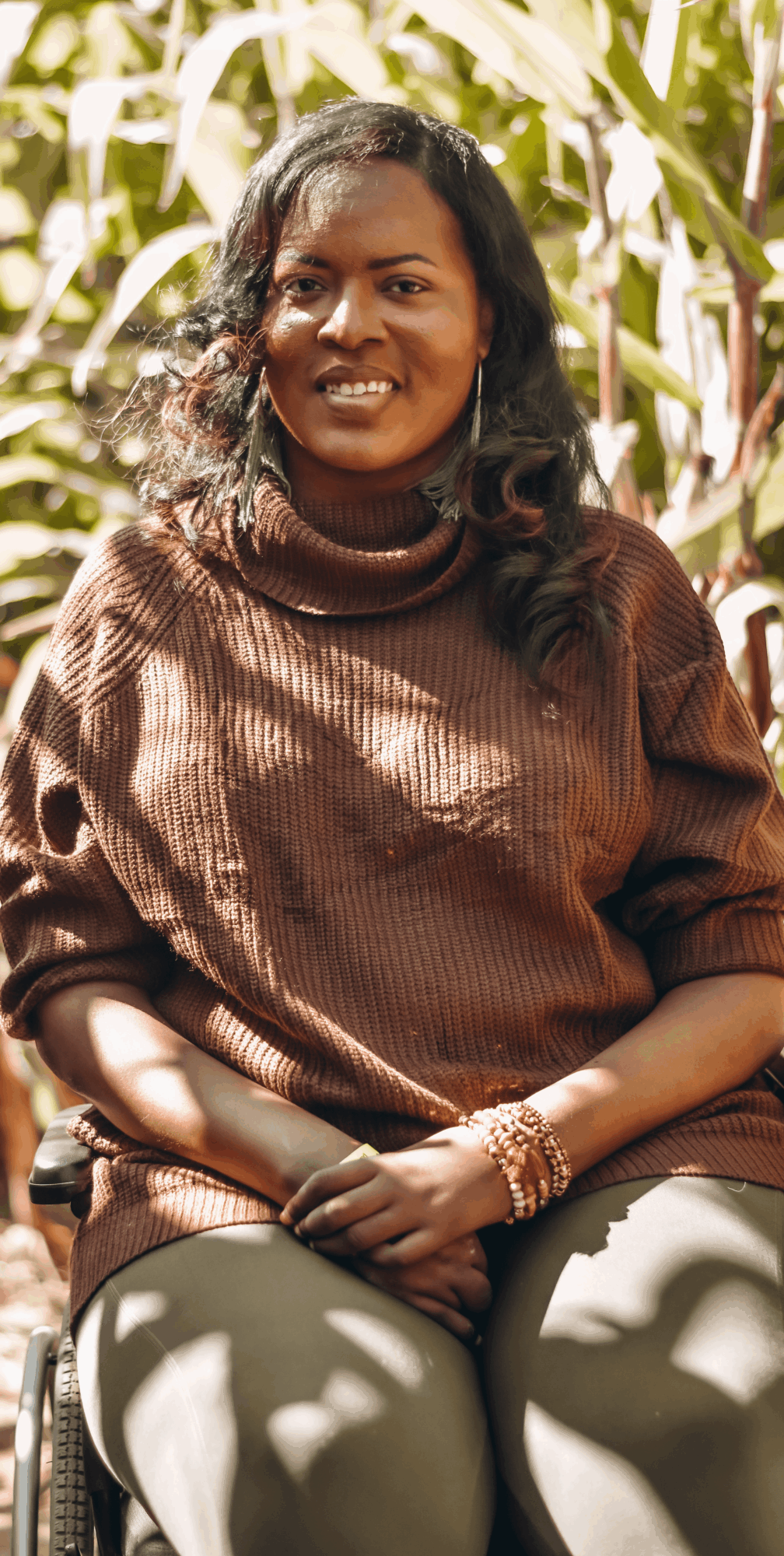 Woman with curly hair smiling outdoors, wearing a brown sweater and sitting with hands on lap, surrounded by plants.