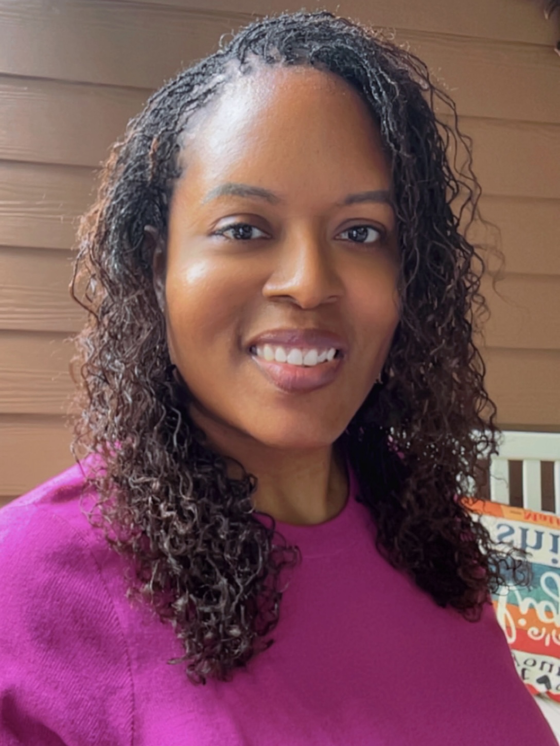 Portrait of a smiling woman with curly hair wearing a purple top, standing indoors with a wooden background.