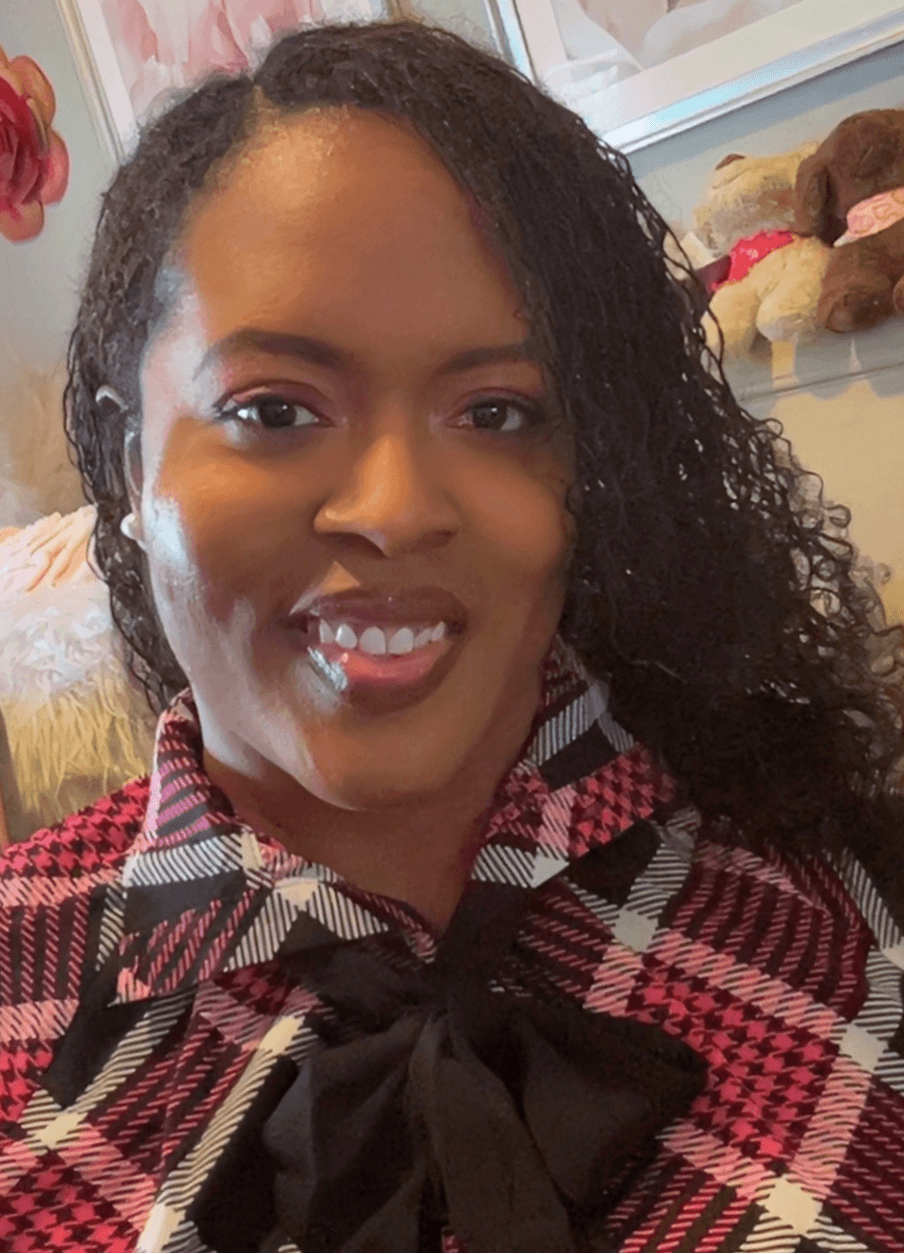 Woman with curly hair smiling, wearing a plaid shirt with a bow, in a room with stuffed animals.