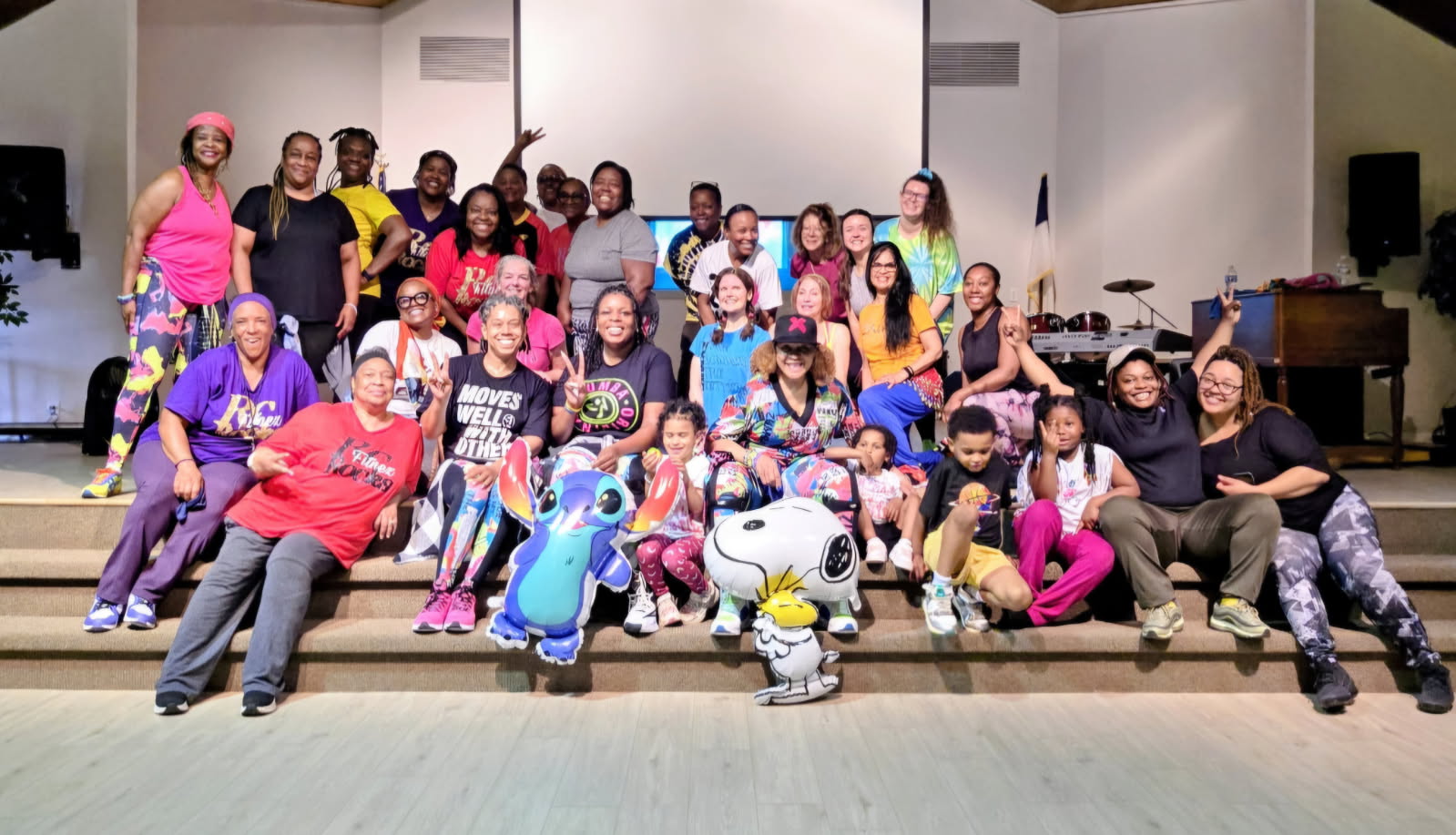 Group of children and adults on stage, some holding balloons and mascot costumes, smiling for a photo.