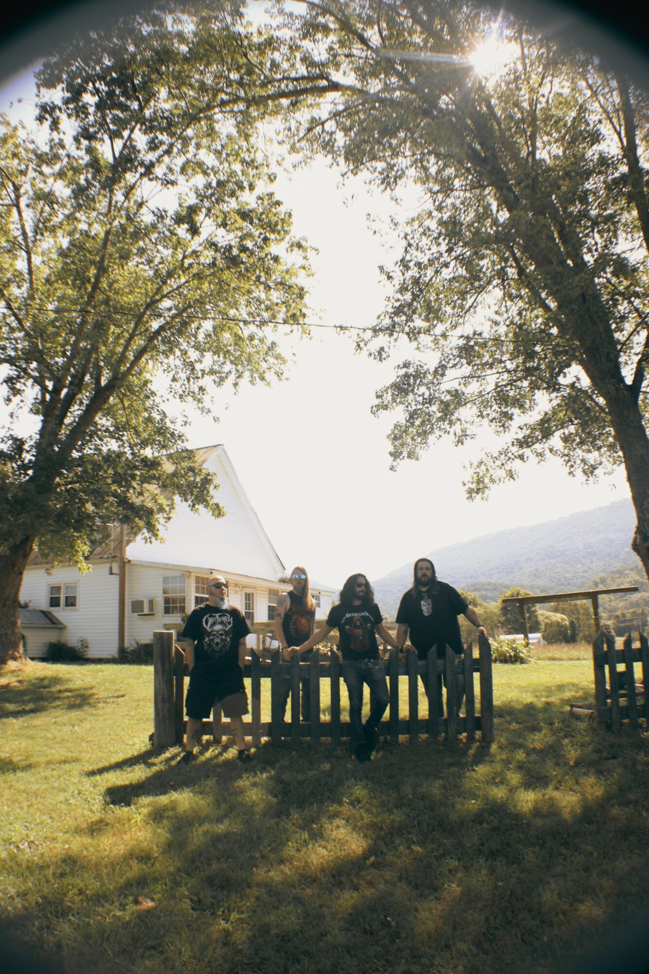 Group of five people sitting on a bench outdoors under trees with sunlight shining through leaves, a house in the background.