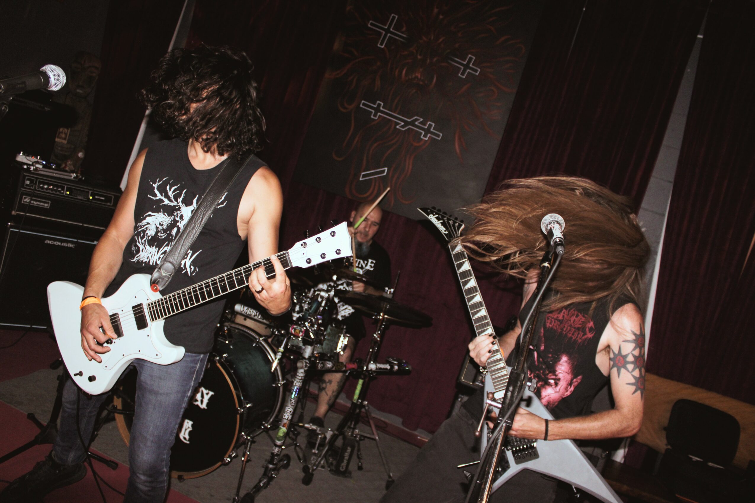 Two musicians perform on stage, one playing guitar and the other with long hair, tattoos, and a guitar, with a drum set behind them.