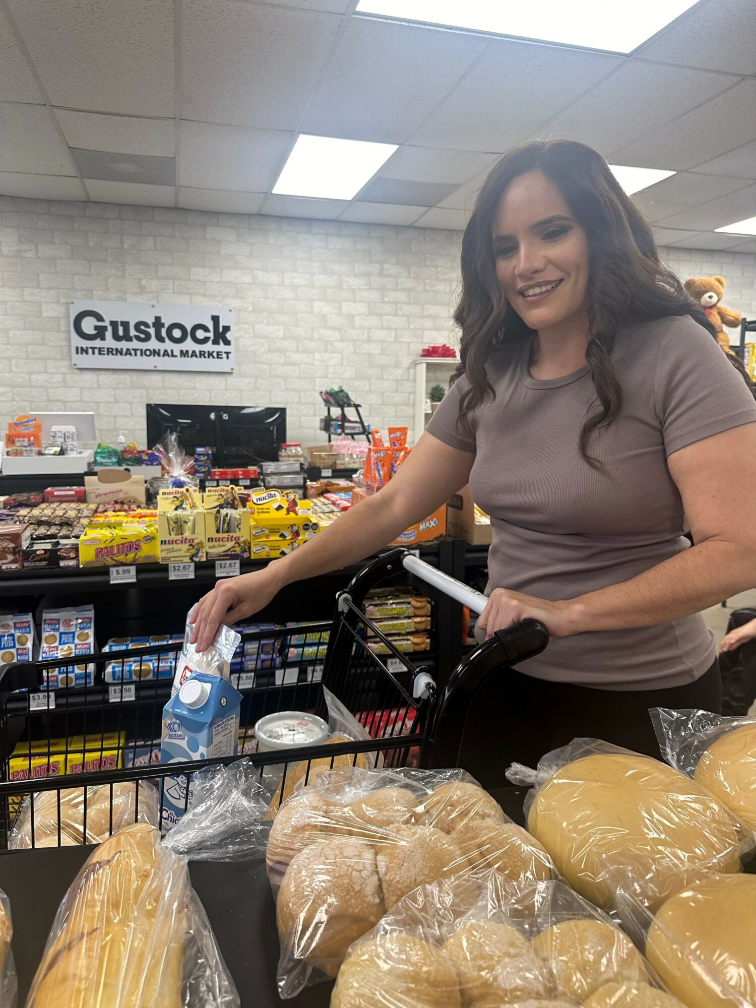 Woman shopping at a grocery store, reaching for items on a shelf, with bread and packaged goods nearby.