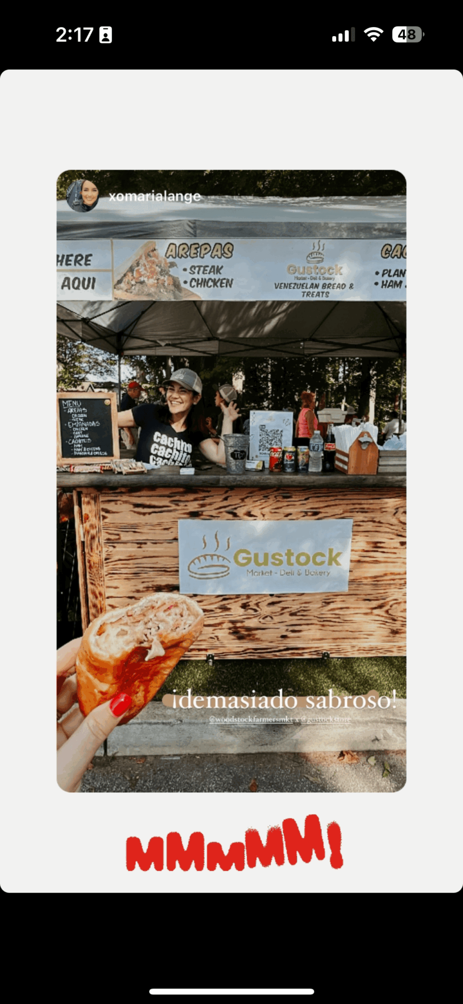 Person holding a drink in front of a food stand with a Gustock sign, outdoor setting, sunny day.