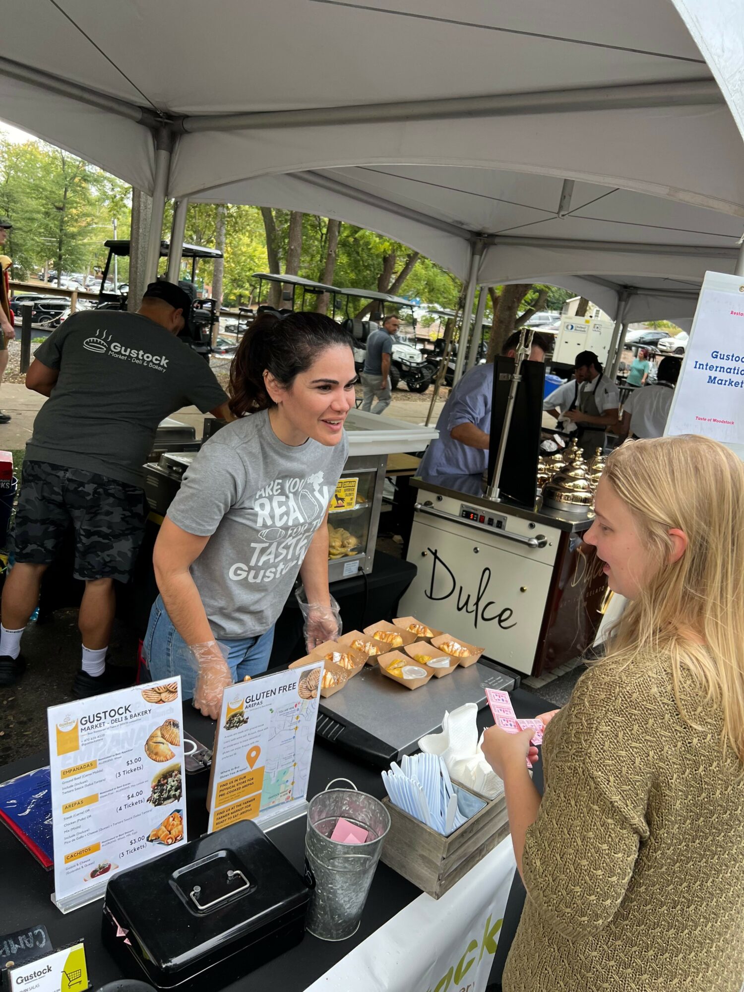 Woman with dark hair and glasses at a food stall, talking to a blonde woman, with menu and food items on table.