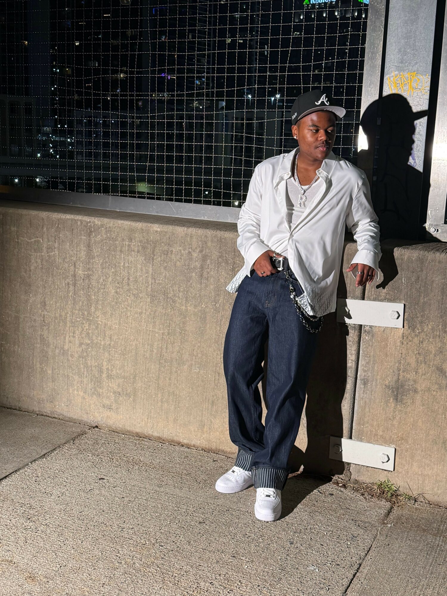 Man standing outdoors near a concrete wall, wearing a white shirt, dark pants, and a cap, with a fence in the background.
