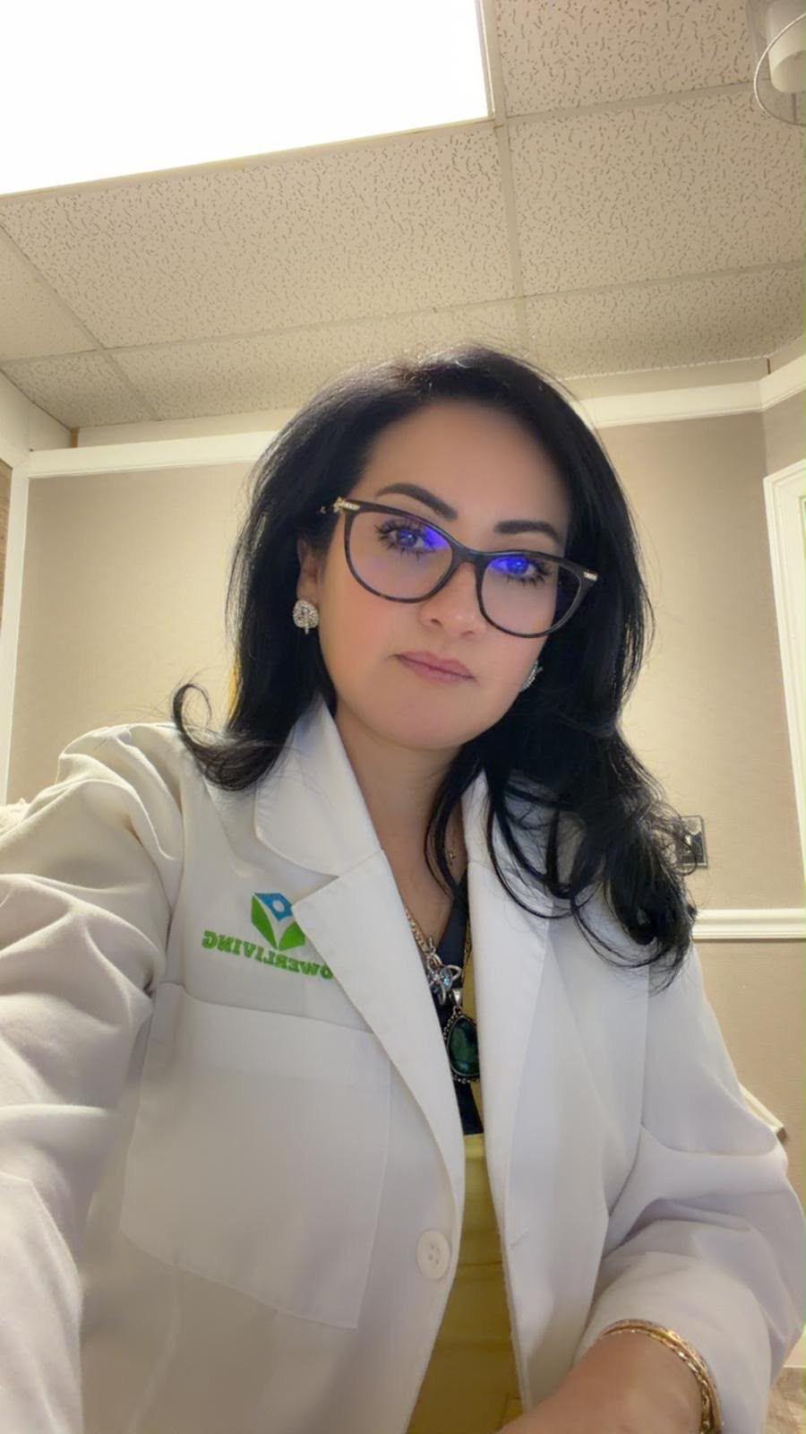 Woman with dark hair and glasses wearing a white coat in an indoor setting.