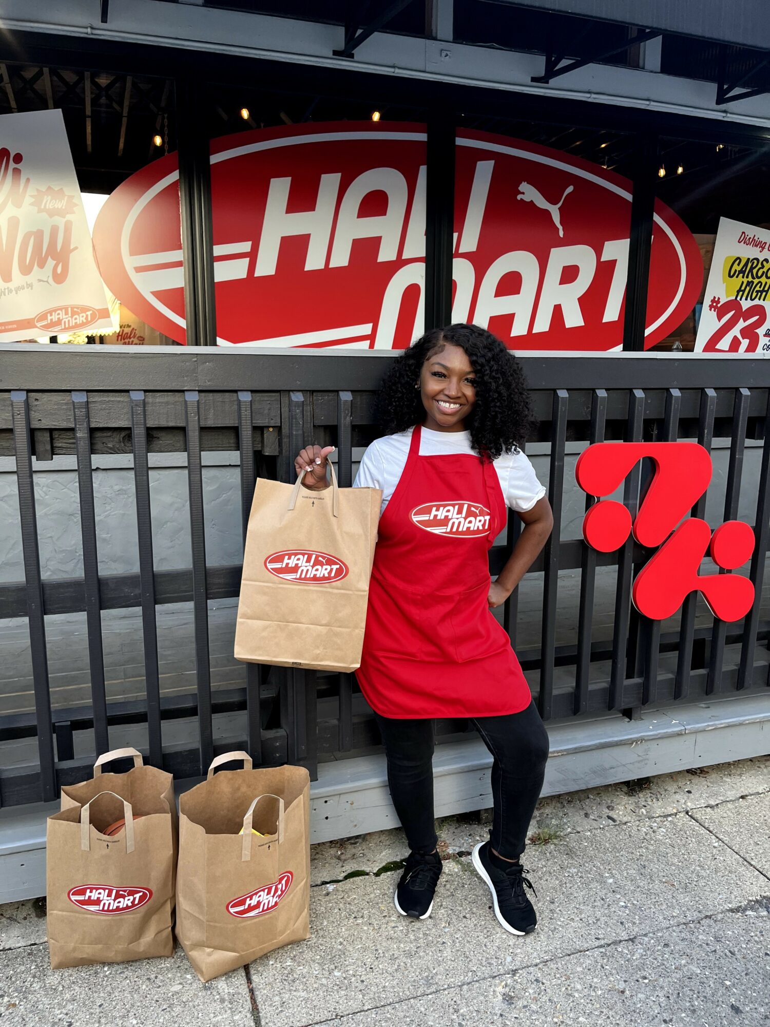 Woman smiling holding a paper bag outside a restaurant with a red and white sign, two paper bags on the ground.