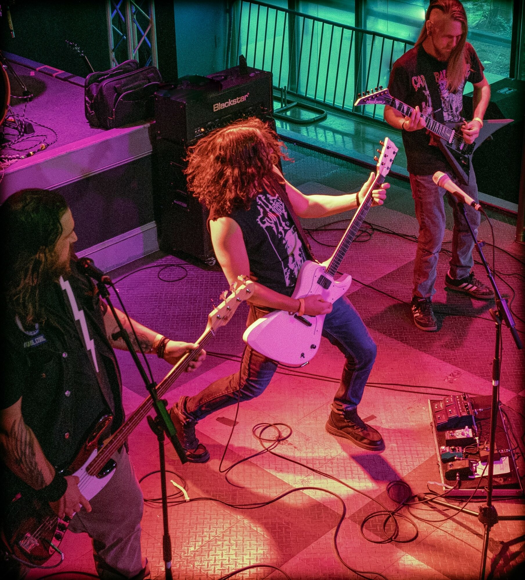 Three musicians perform on stage with guitars, one with curly hair, in a colorful setting.