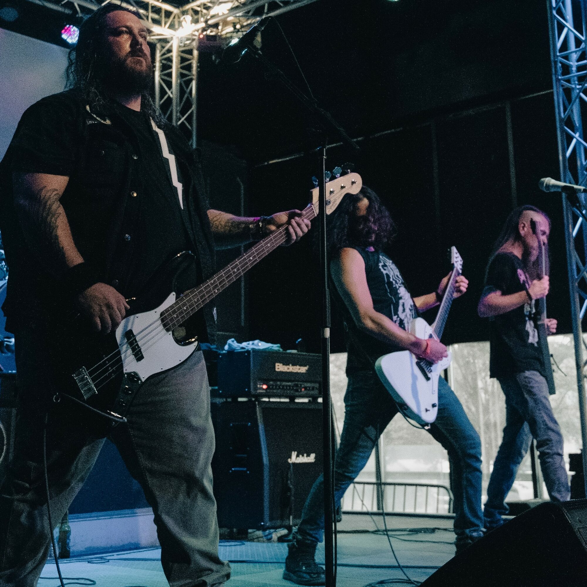 Three musicians perform on stage with guitars and microphones, stage equipment visible in the background.