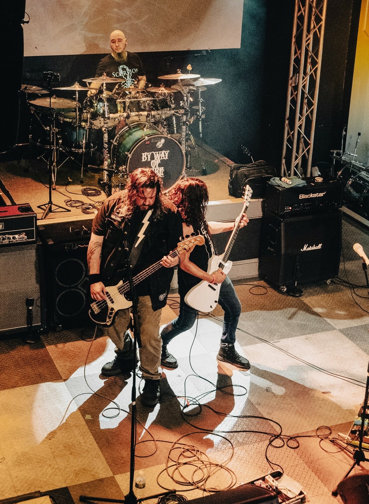 Two guitarists perform on stage with a drummer in the background, stage equipment, and lighting visible.