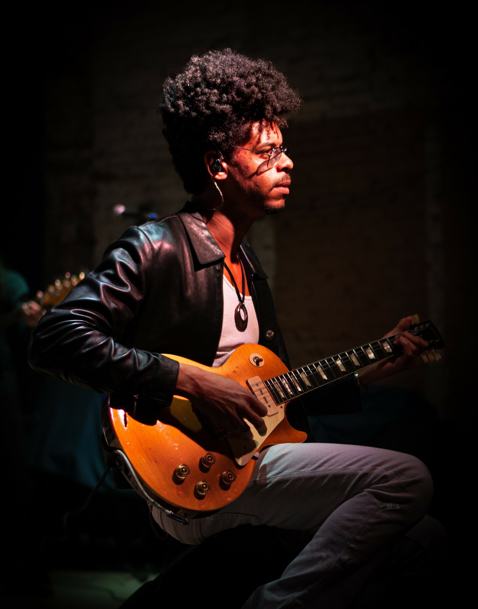 Person with curly hair playing an orange electric guitar, wearing glasses and a leather jacket, sitting in a dark setting.