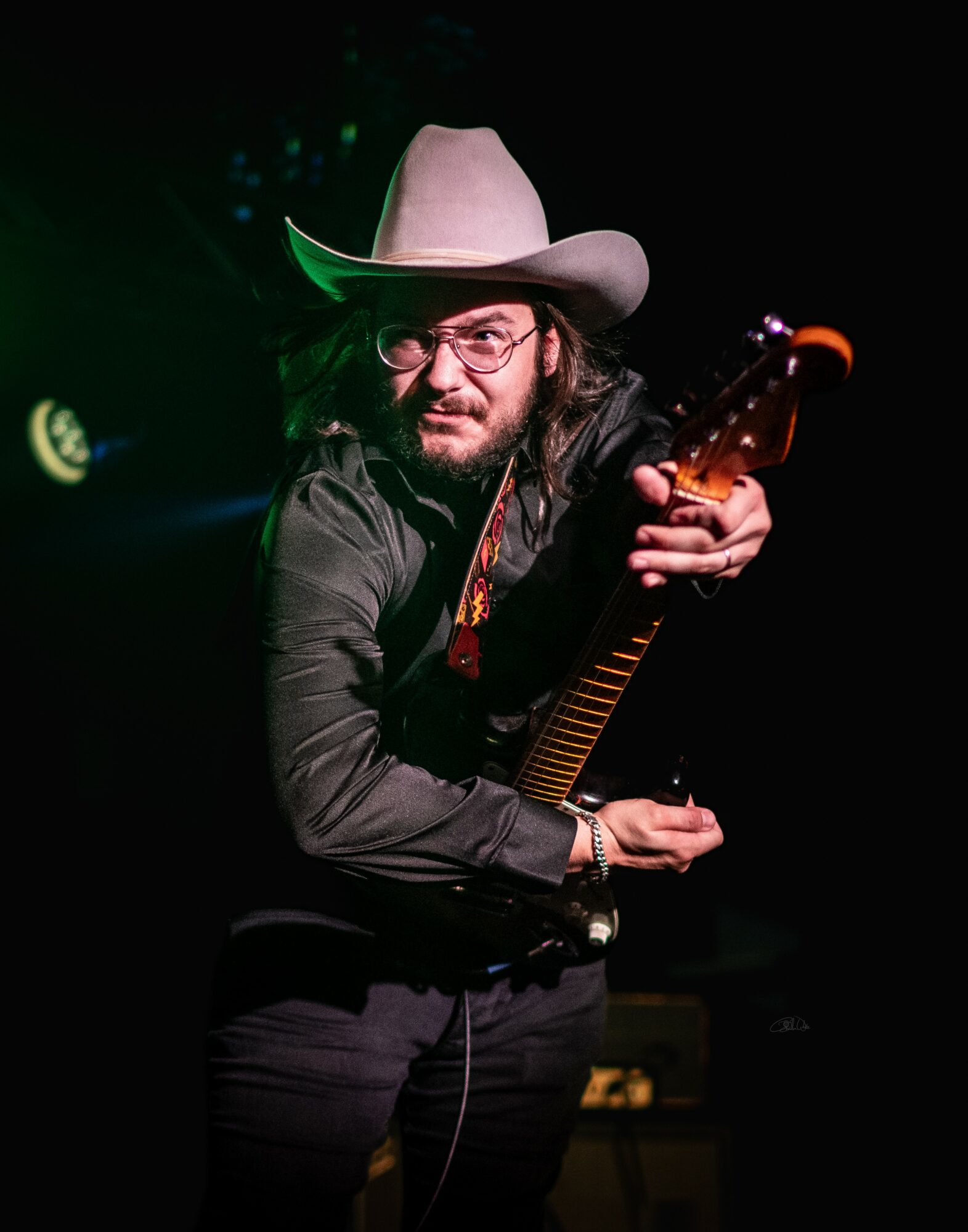 Person wearing a cowboy hat and glasses, holding a guitar, in a dark setting with stage lights.