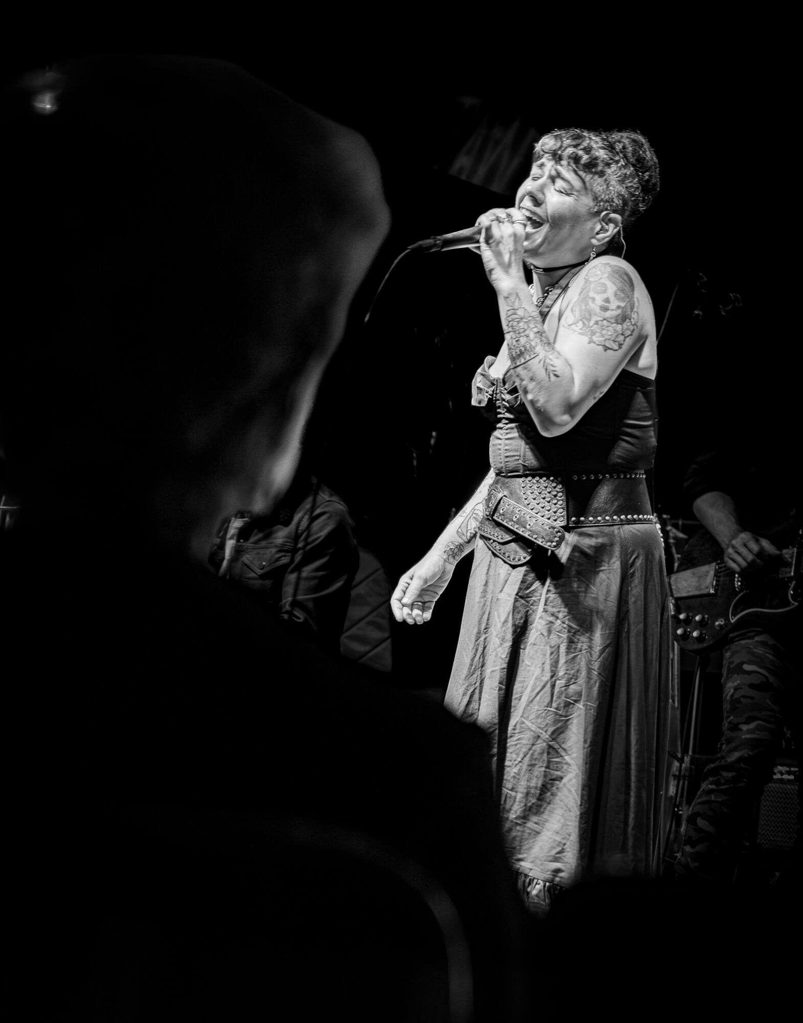 Performer singing into a microphone on stage, with tattoos visible, in black and white.