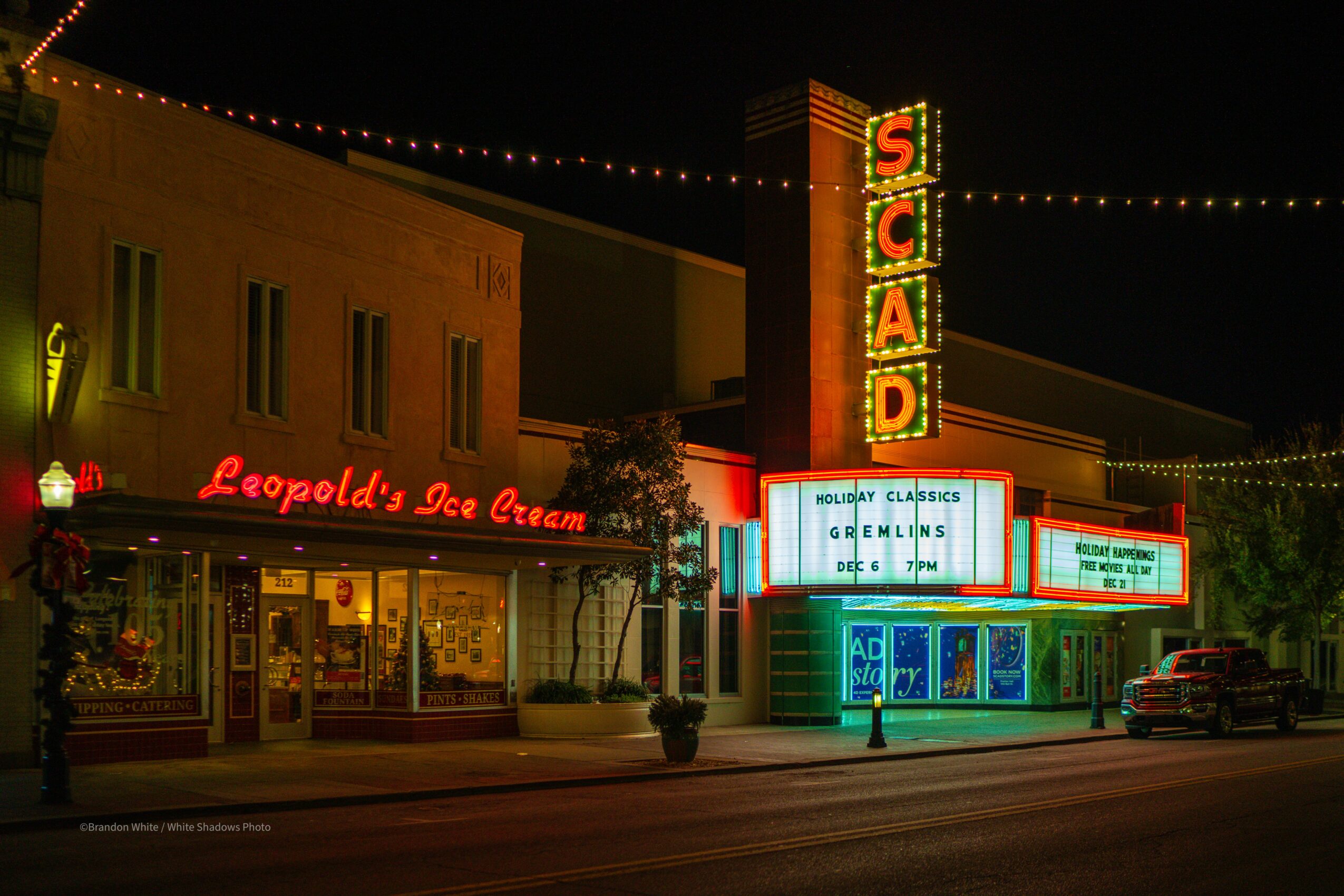 Night scene of a city street with illuminated signs for Leopold's Ice Cream and a theater marquee, with a parked car and string lights.