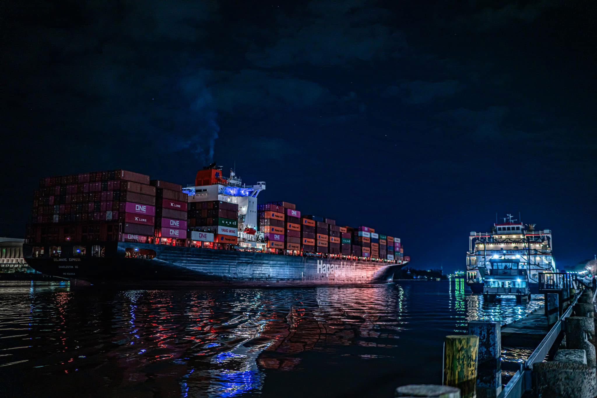 Large cargo ship with containers at night, reflecting on water, with illuminated buildings nearby under dark sky.