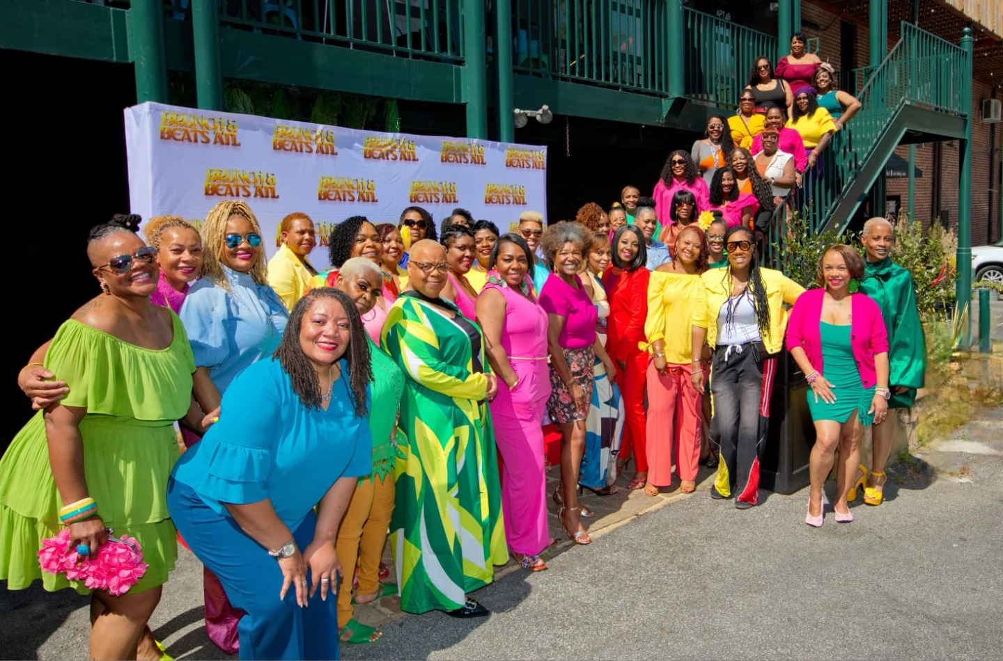 Group of women and children in colorful clothing standing outdoors, some on stairs, in front of a backdrop with text.