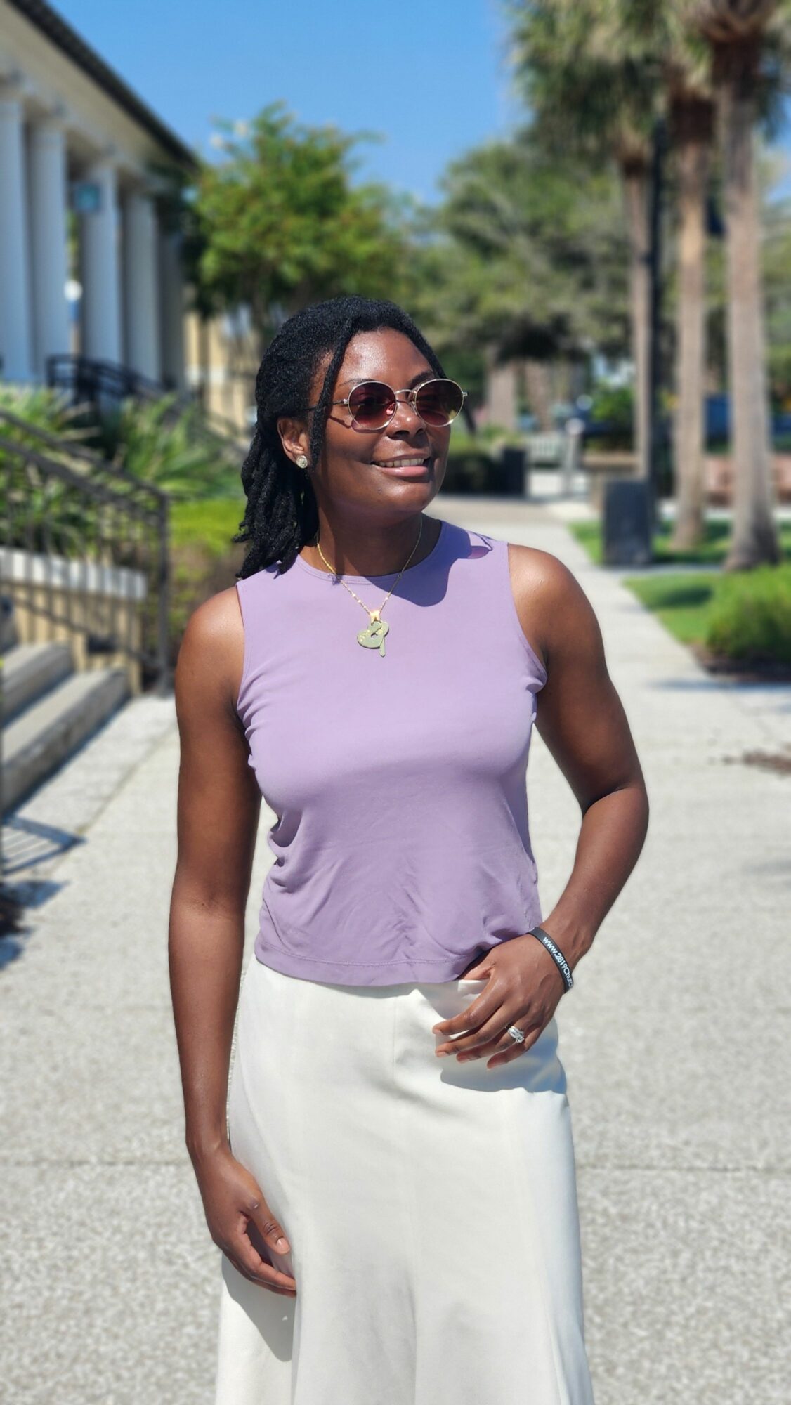 Woman wearing sunglasses, purple top, and white skirt standing outdoors on a sunny day, smiling, with trees and buildings in background.