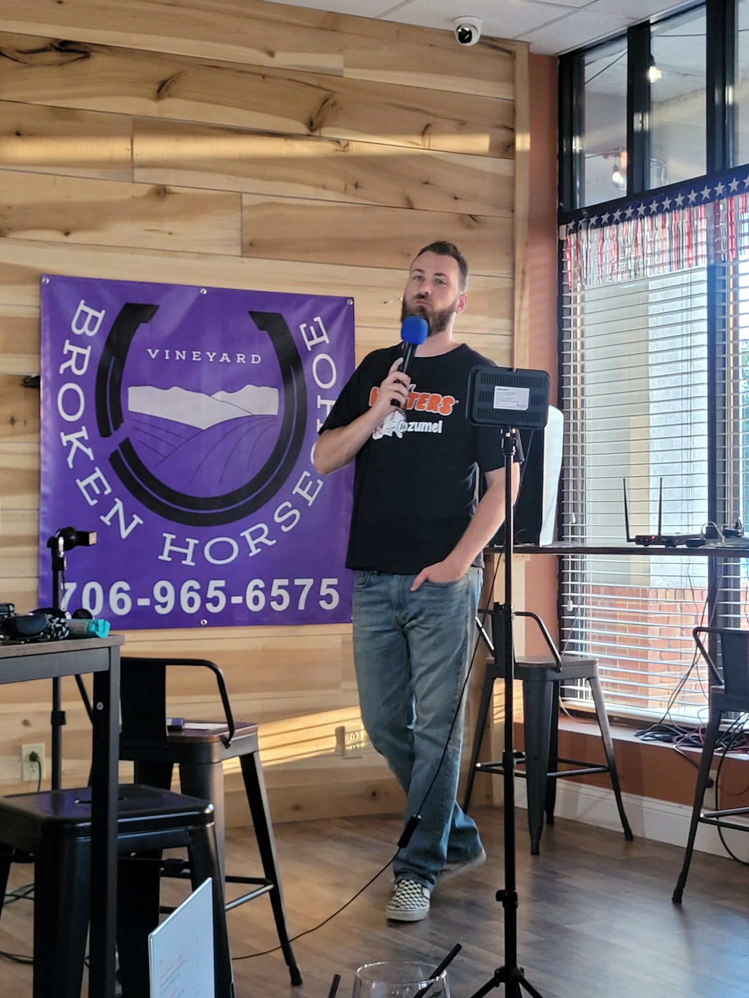 Man with a beard speaking into a microphone in a room with wooden walls and large windows, a purple banner behind him.