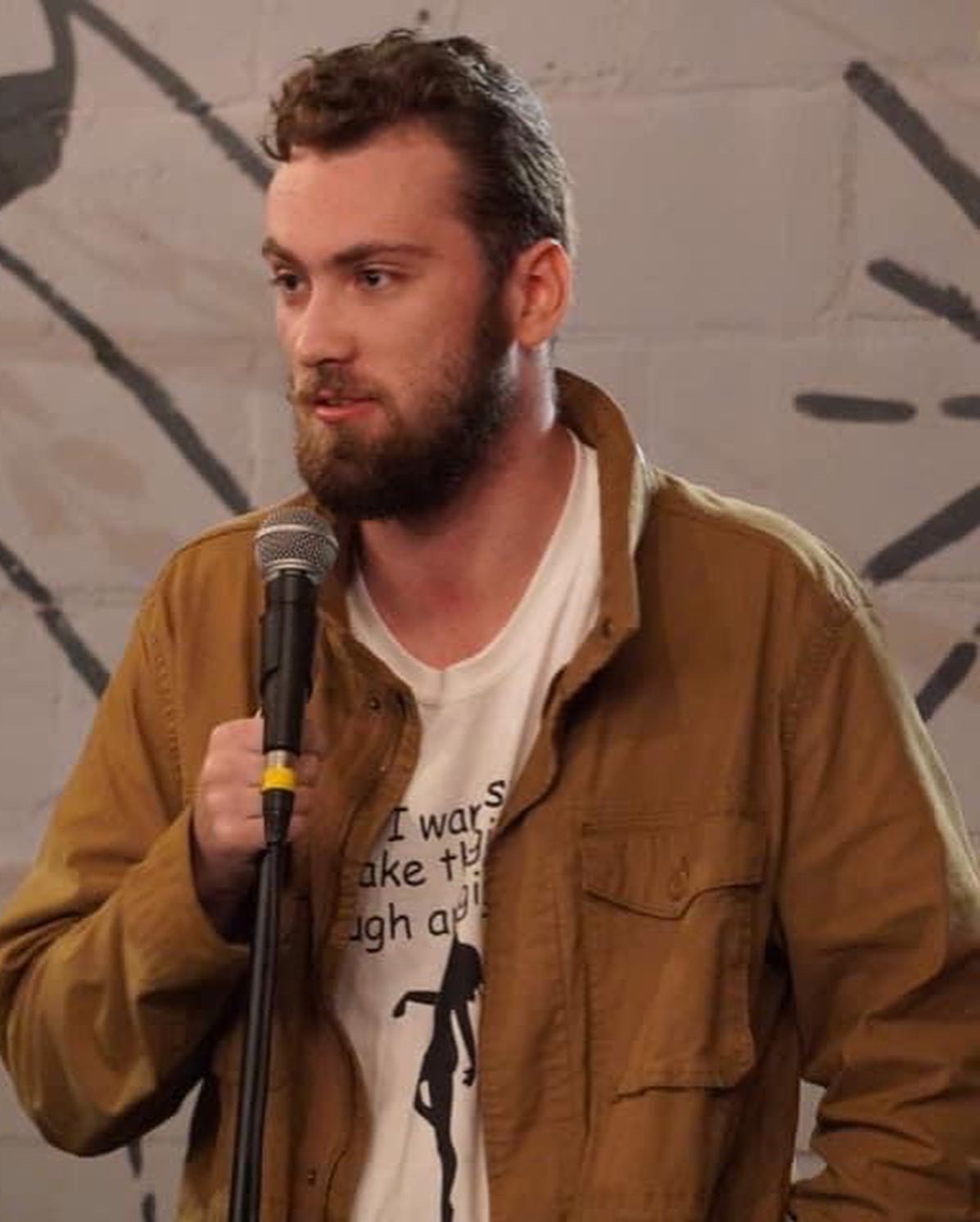 Man with beard holding microphone, wearing brown jacket and white shirt, standing against a wall with graffiti.