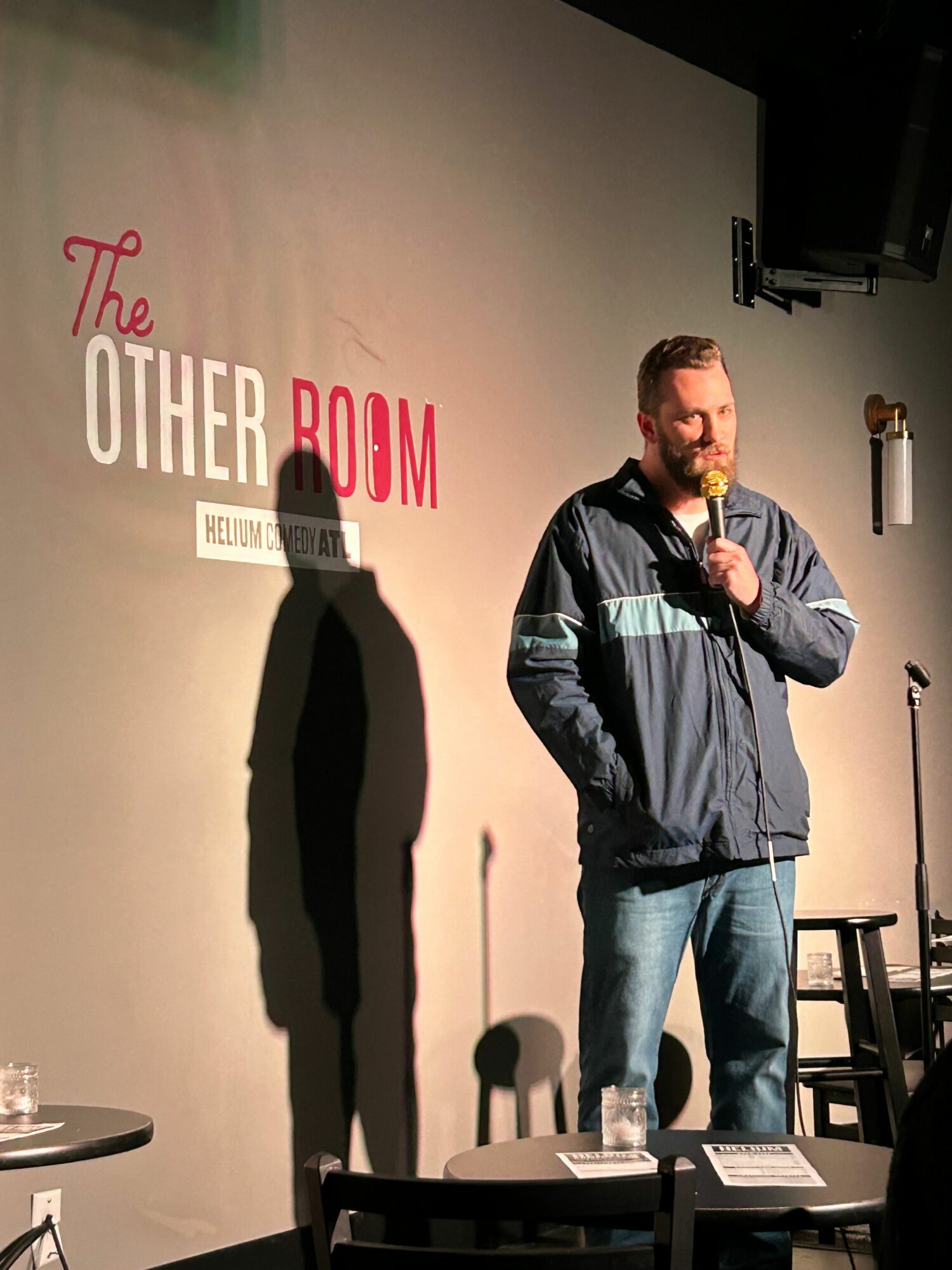 Man standing on stage with microphone, shadow cast on wall, behind a table, in front of a wall with red text.