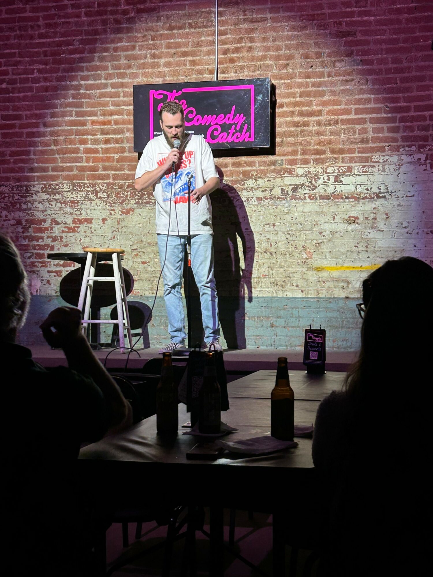 Person standing on stage with microphone, brick wall background, pink neon sign, audience silhouette, and a table with bottles.