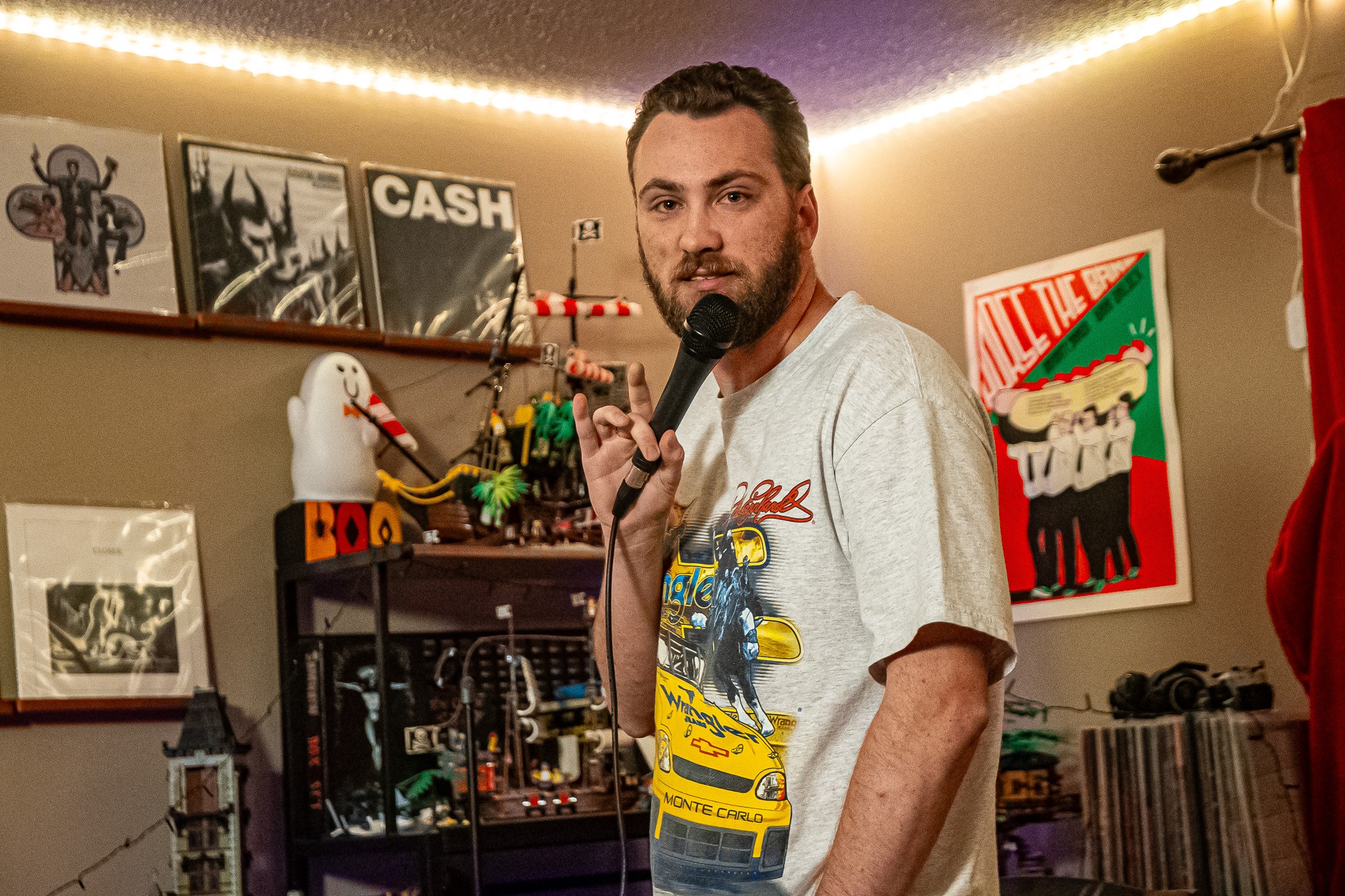 Man with beard holding microphone in a room decorated with posters and toys, looking at the camera.