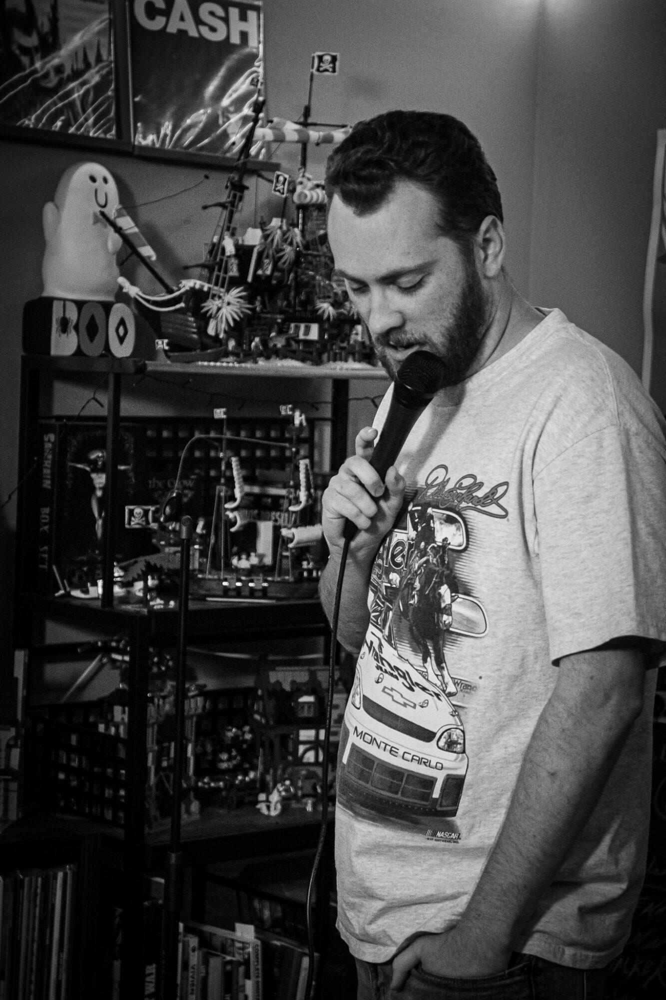 Man with beard holding microphone, looking down, in room with electronic equipment and a plush toy on shelf.