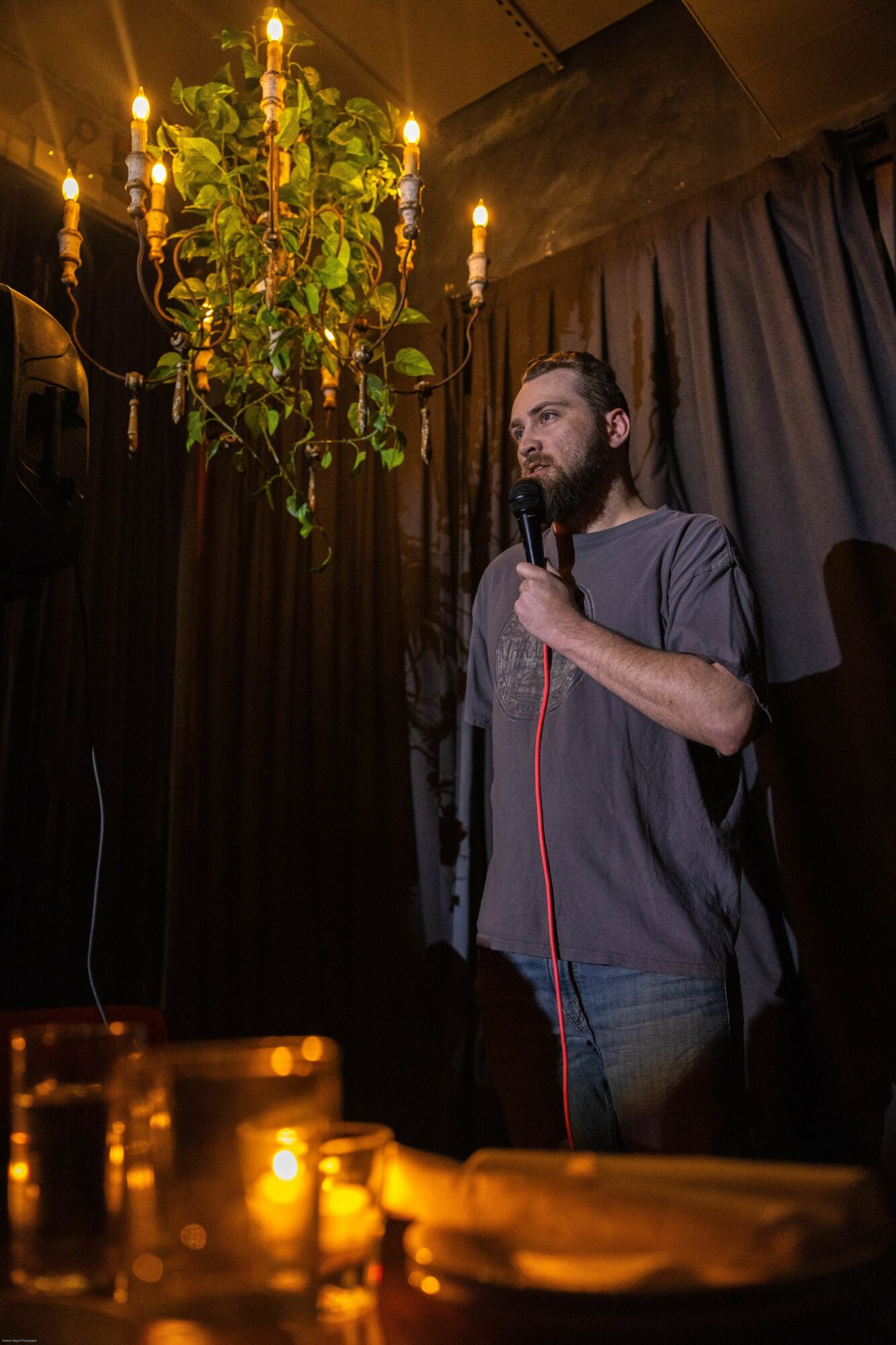 Man with a beard holding a microphone on stage, black curtains behind, chandelier overhead, candles on table in foreground.