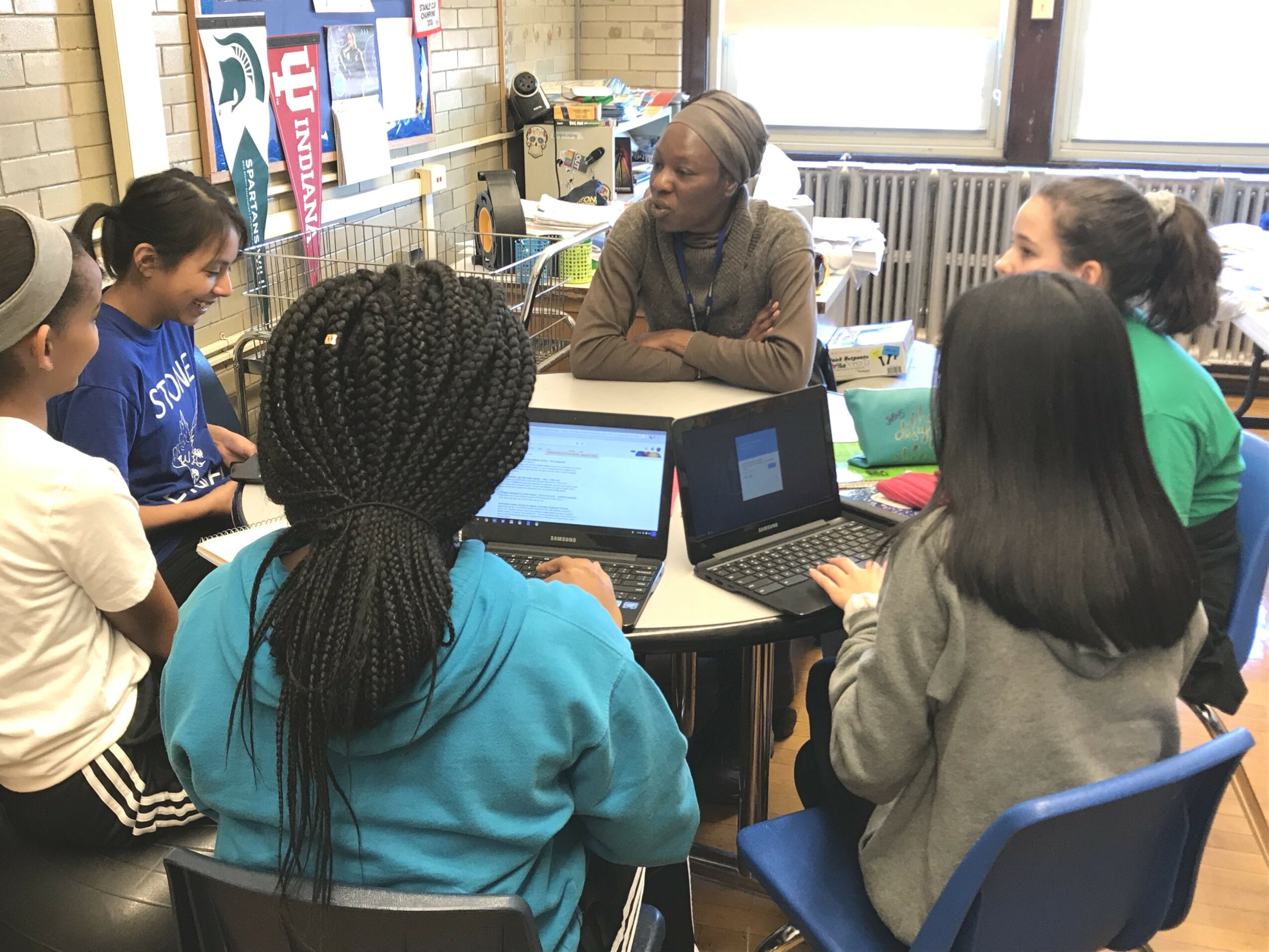 Group of people sitting around a table in a classroom, engaging in discussion with laptops open.