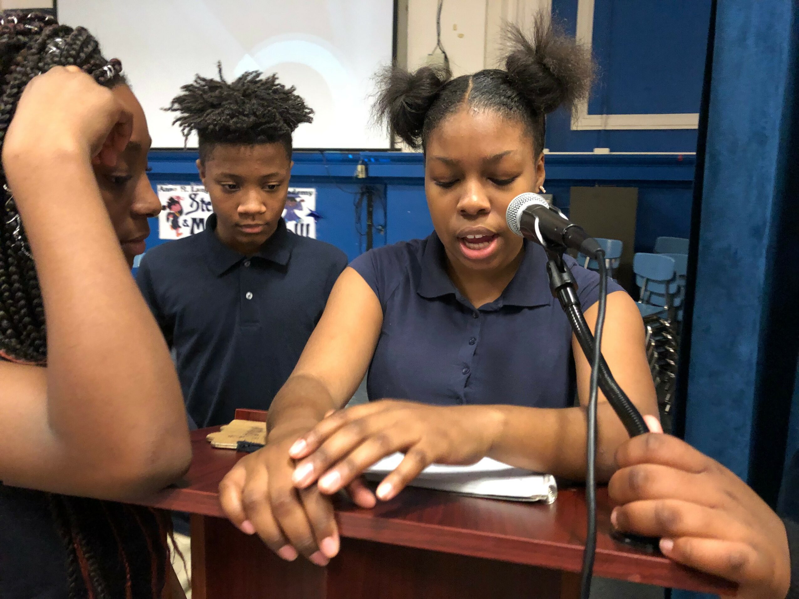 Two girls and a boy are at a table, with one girl speaking into a microphone, others listening.