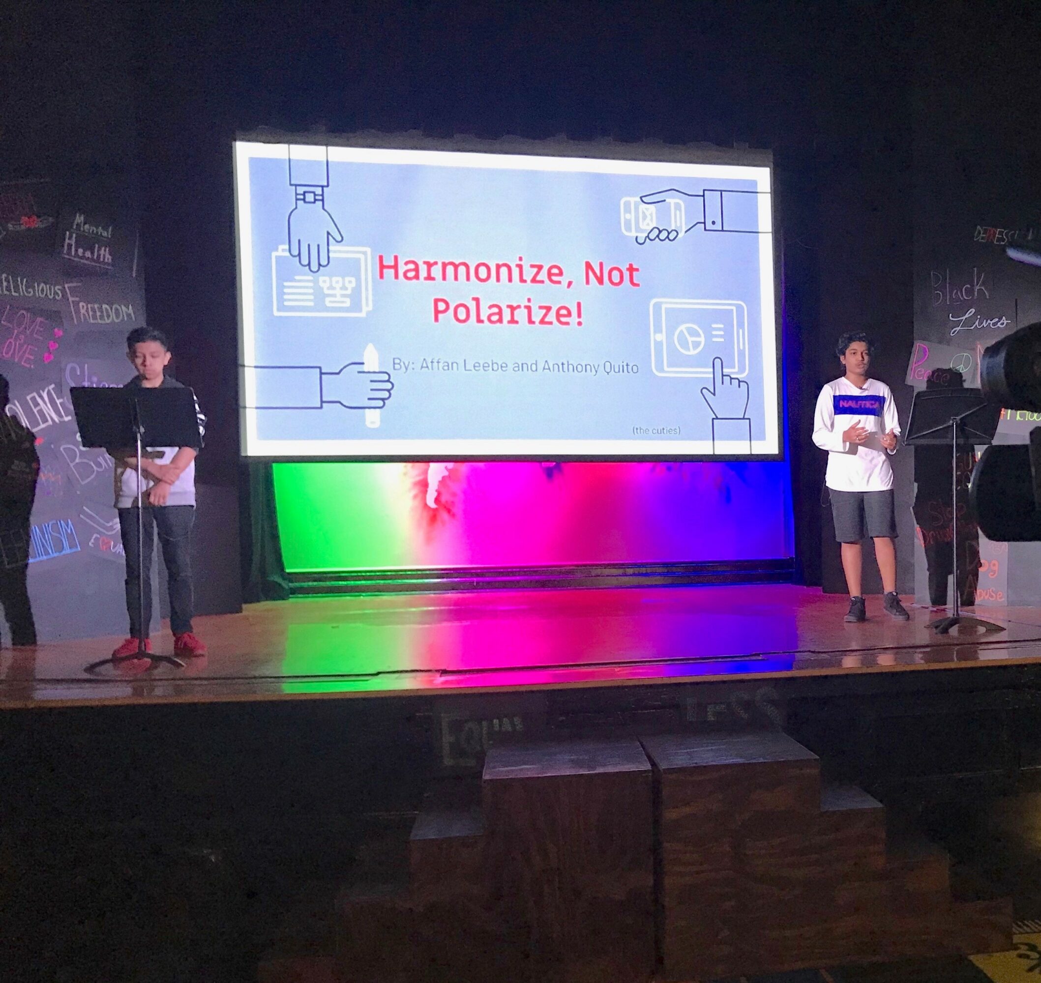 Three people on stage in front of a large screen displaying a presentation titled 'Harmonize, Not Polarize!'.