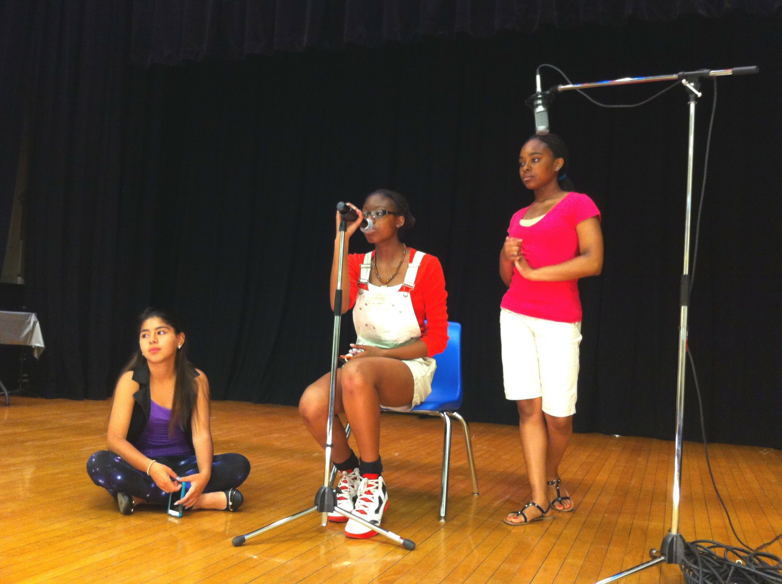 Three girls on stage, one sitting cross-legged, one standing at microphone, and one standing nearby, with a black curtain background.