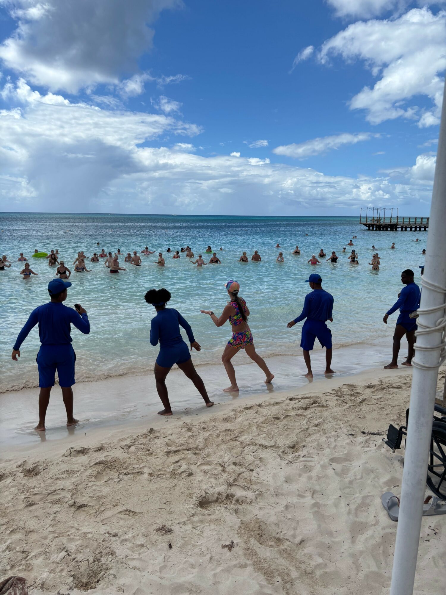 People walking on sandy beach near water with boats and a pier in the background, under a partly cloudy sky.