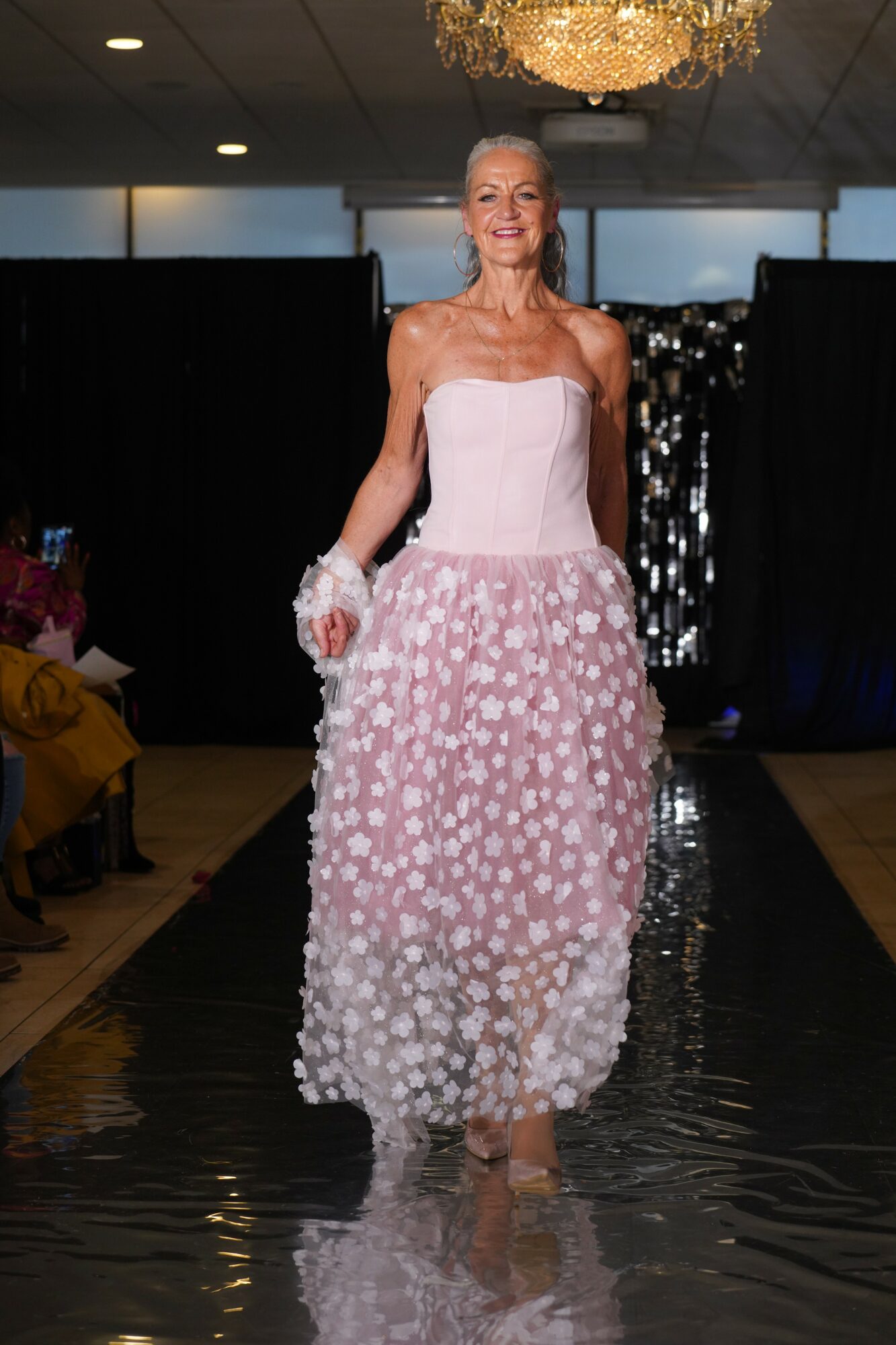 Woman walking on runway in pink strapless dress with floral pattern, holding a bouquet, smiling, chandelier overhead.