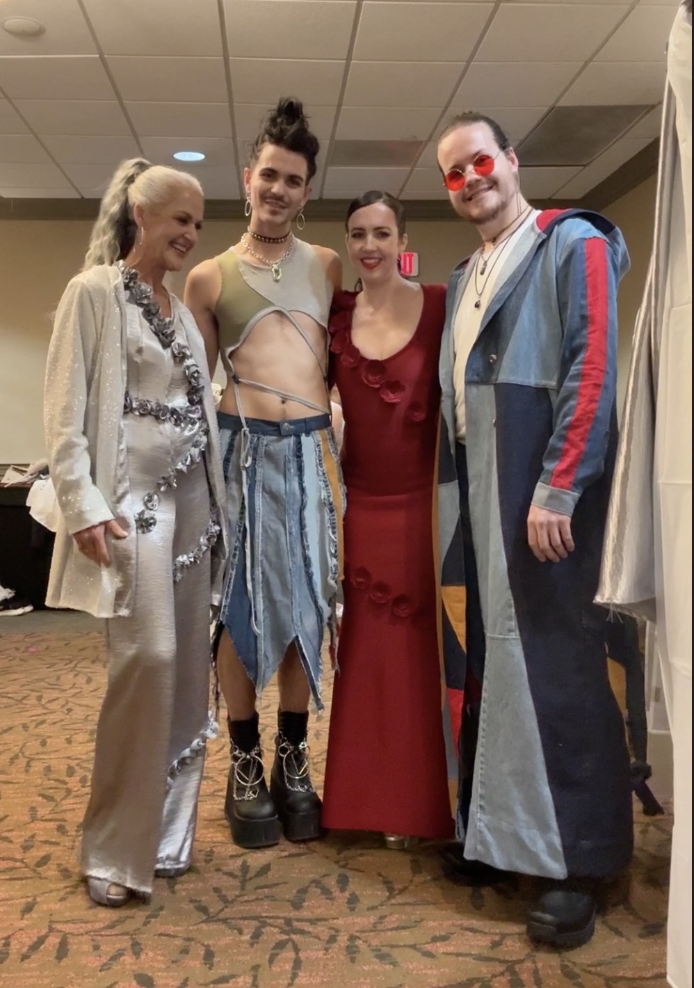 Four people in costumes standing together indoors, smiling, with a patterned carpet and ceiling tiles visible.